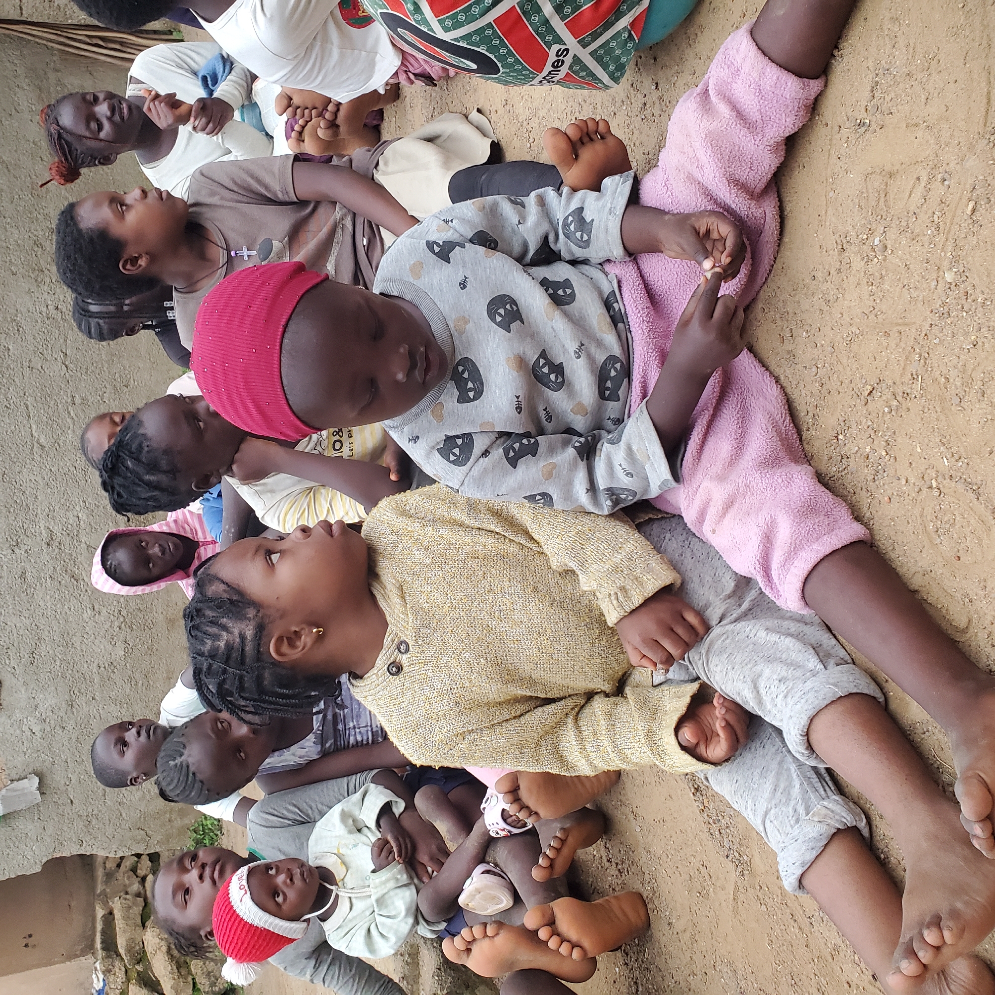 In an outdoor environment with a dirt ground and a rough concrete wall in the background, a group of Black children are seated on the ground. The children, ranging in age from toddler to pre-teen, are dressed in casual clothing. The foreground is dominated by two young girls. The girl on the left wears a yellow knitted sweater and grey pants, while the girl on the right is in a grey long-sleeved shirt with a cat and fish pattern and pink pants. Both girls are looking downwards. Other children are visible in the background, some looking towards the camera, others engaged in conversation. One toddler in a red hat and a light-colored outfit sits in the middle ground. The scene appears to be a candid moment captured in a rural or less urbanized setting.