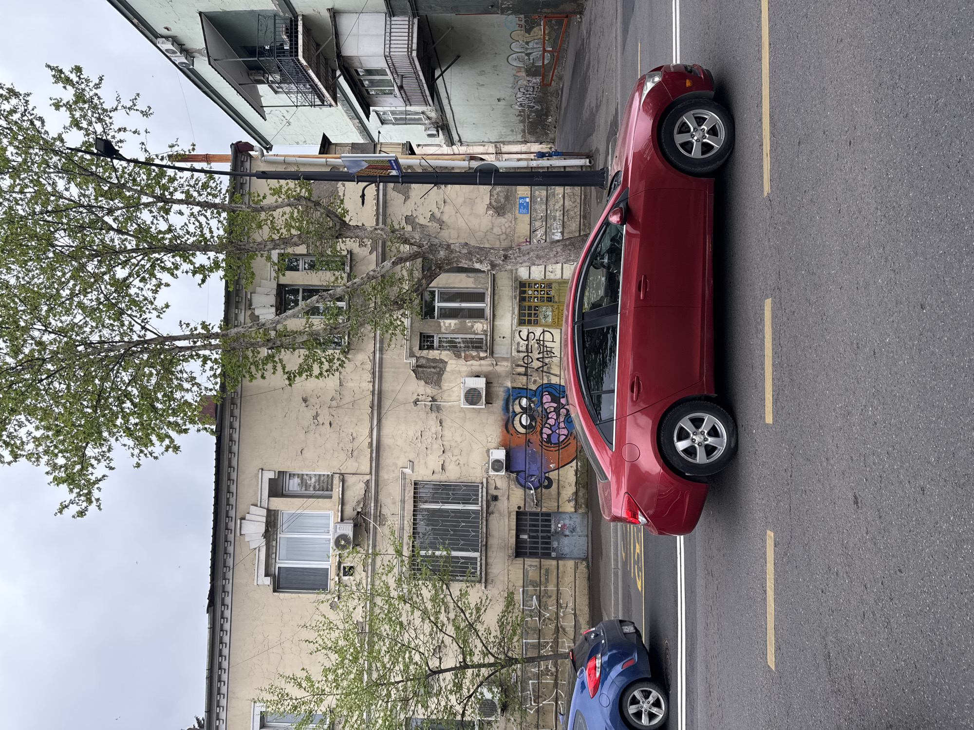 The image depicts a street in an urban setting in Unknown, Unknown. A red sedan is parked along the side of the road next to a building with peeling paint and graffiti. A blue car is parked behind the red car. A tree is visible in front of the building, and a lamppost stands nearby. The road is marked with yellow lines. The sky is overcast.