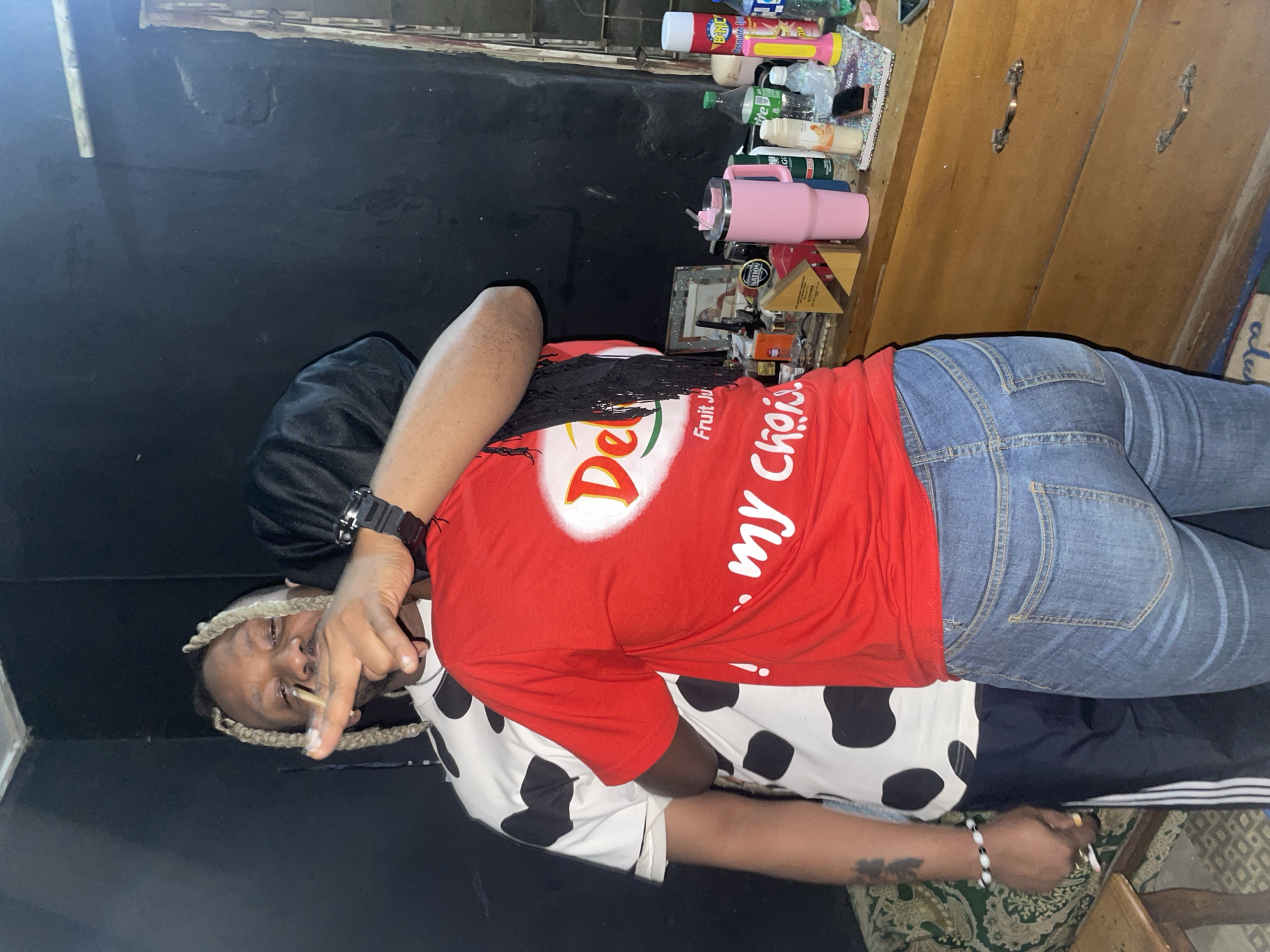 The image shows a person in a room. The person wears blue jeans, a red t-shirt with a white logo and text, and a white and black spotted garment. The person has braided hair and is wearing a wristwatch on the left wrist. They are holding something between their fingers. A wooden cabinet with objects on top is visible in the background. The room has a dark wall. It is unknown whether the image was taken in Jos, Nigeria.