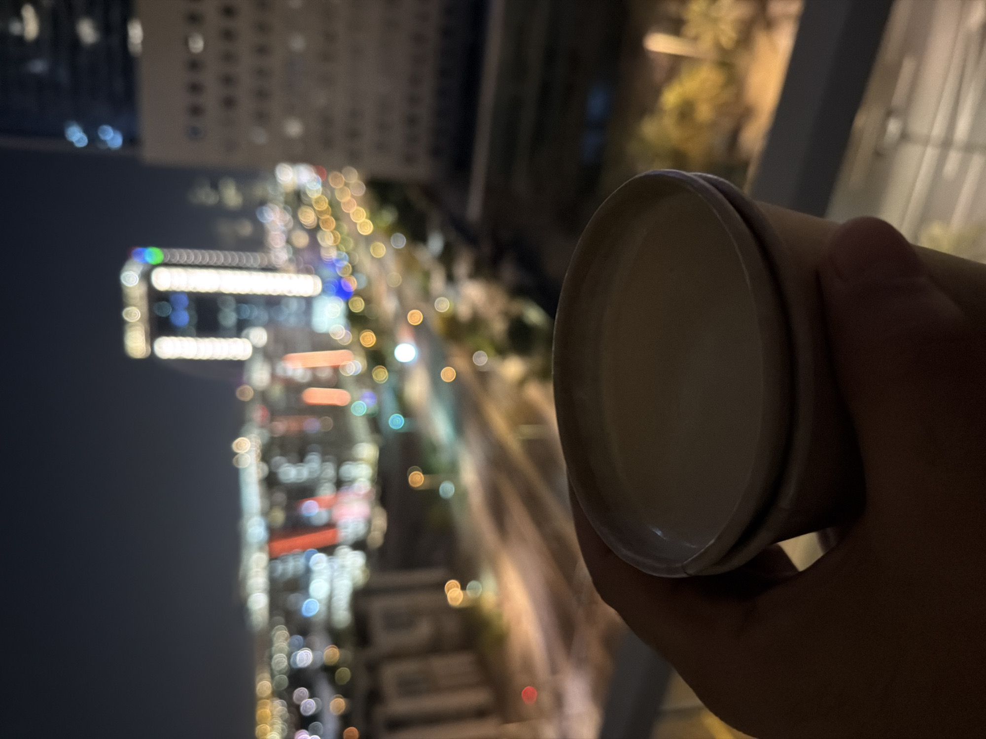 A hand holds a light-colored paper cup of coffee in the foreground. The cup is positioned in the lower right portion of the frame, obscuring part of the background. The background depicts a blurred cityscape at night. Numerous lights from buildings and streetlights create a bokeh effect, with circular and elongated shapes of varying colors, including white, yellow, blue, and red. One prominent building in the mid-ground is brightly illuminated with vertical white lights and a green light at its apex. The scene appears to be taken from an elevated vantage point, likely a balcony or window, overlooking a street or intersection. The sky is dark, suggesting nighttime.
