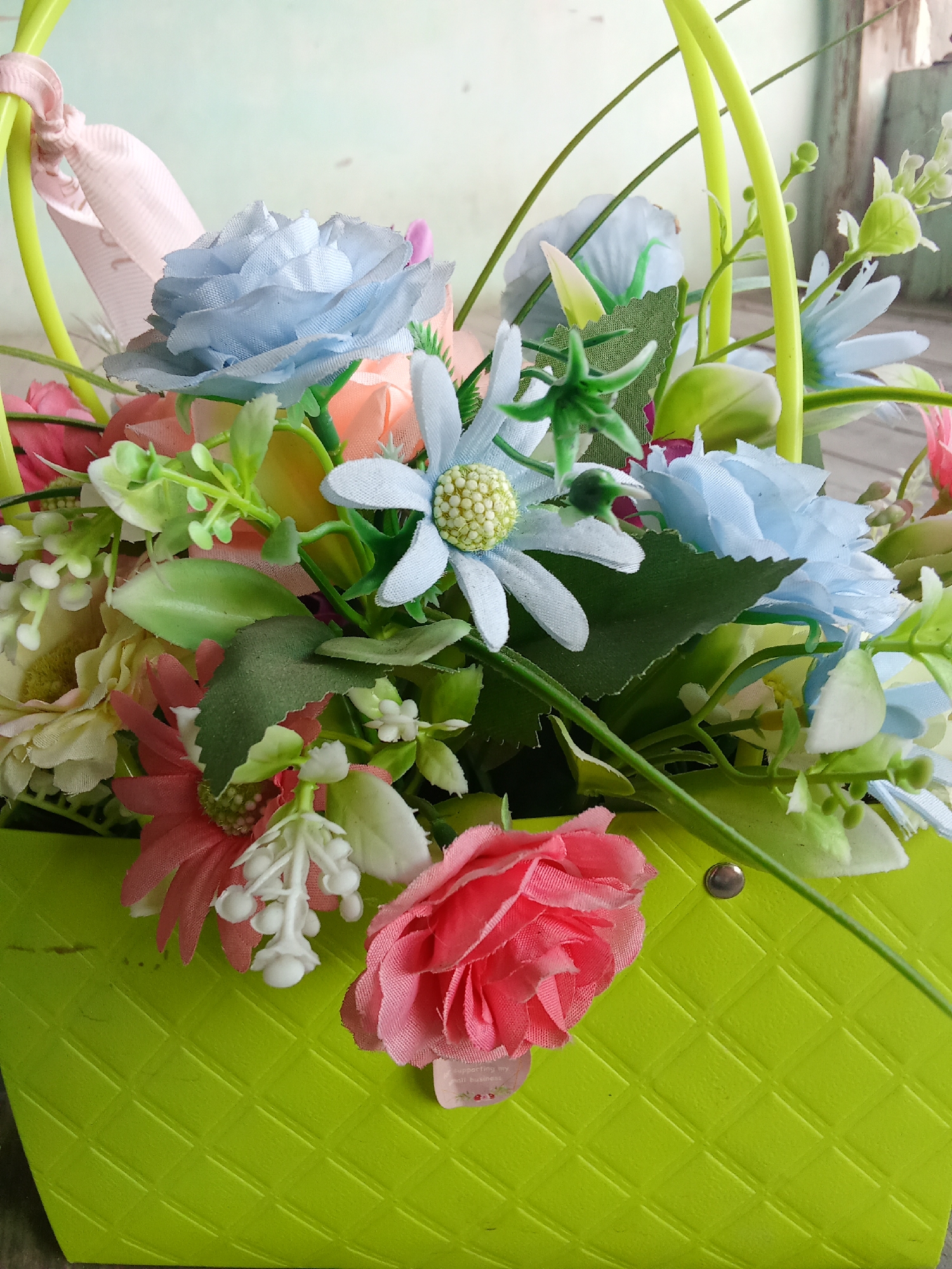 A close-up, slightly high-angle shot shows a vibrant arrangement of artificial flowers spilling out of a bright lime green basket. The basket has a textured, diamond-patterned surface and a thin, light pink ribbon tied to its green handle.
The floral arrangement is dense and colorful, featuring a variety of blooms in shades of pink, light blue, cream, and peach. Prominent among the flowers are several large, layered pink roses, delicate light blue roses with intricate petal details, and smaller clusters of white flowers resembling baby's breath. Green leaves and artificial greenery are interspersed throughout the bouquet, adding depth and fullness.
In the background, a light blue-green wall is visible, with a hint of a wooden structure to the right. The lighting suggests an indoor setting, possibly on a surface that is out of focus.
The image is focused on the floral arrangement and the basket, with the background softly blurred. The overall impression is one of cheerful and artificial beauty. There are no people or animals present in the scene. The location of Fergana City, Uzbekistan, is not visually discernible from the provided image.