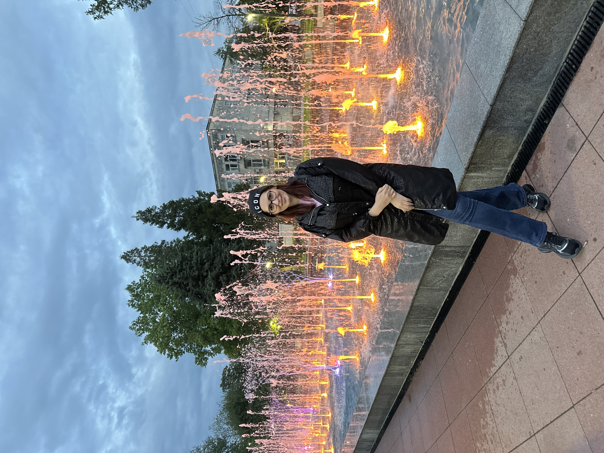 A woman is standing near a fountain. The fountain has many vertical water jets lit with orange and purple lights. The woman is wearing a black jacket, jeans, and a black beanie with white lettering. She is wearing glasses and smiling. Behind the fountain is a building and some trees. The sky is cloudy. Based on the limited information, the specific location in Montana, Bulgaria cannot be determined.