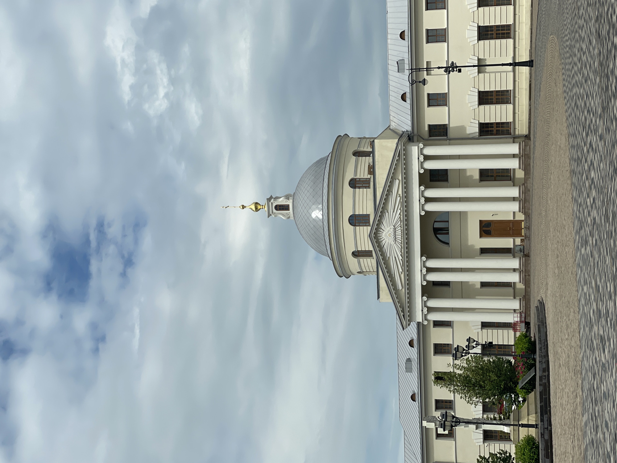 The image captures a daytime view of the Kazan Kremlin's Temple of the Assumption of the Blessed Virgin Mary, also known as the Cathedral of the Annunciation. The building features a neoclassical architectural style, characterized by its prominent portico with classical columns and a large, silver dome topped with a golden cross. The exterior walls are painted a light yellow, contrasting with the grey metal roof and the surrounding cobblestone plaza.
In the foreground, the plaza is paved with geometric patterns of light and dark grey cobblestones, forming a circular area in front of the building. Several black lampposts with multiple globes are strategically placed around the plaza, one of which is located on the right side of the frame. To the left, a small green area with bushes and flowering plants adds a touch of nature to the urban landscape. A tree stands near the center-left of the plaza.
The sky above is overcast with thick, grey clouds, suggesting a cloudy day. The overall scene is static, with no people or animals visible. The image is taken from a slightly elevated perspective, looking towards the main facade of the temple. The lighting is diffused due to the clouds, resulting in soft shadows. The architectural details, such as the windows, cornices, and decorative elements above the windows, are clearly visible.