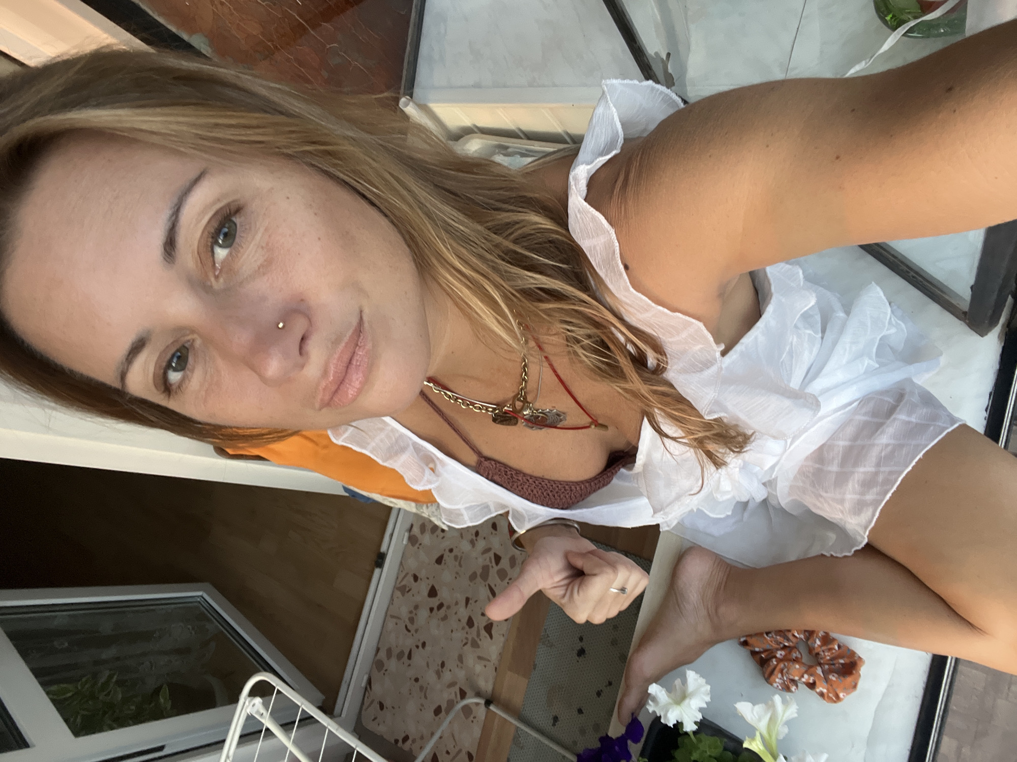 An overhead, high-angle medium shot captures a Caucasian woman from the chest down, sitting indoors on a balcony in Samara, Russia. She is wearing a brown, crocheted bikini top and a white, ruffled dress that exposes her legs and arms. Her right hand is raised, giving a thumbs-up gesture, and her left foot is visible, positioned over her right thigh. A gold necklace with pendants adorns her neck, and a red thread is visible around her neck. A scrunchie with an orange and brown floral pattern lies on a white surface next to her left leg. To the left of the frame, purple and white petunias are visible, suggesting a plant on the balcony. The background shows a wooden floor and a partially open glass door leading outdoors. A white drying rack is also visible on the left side of the image.