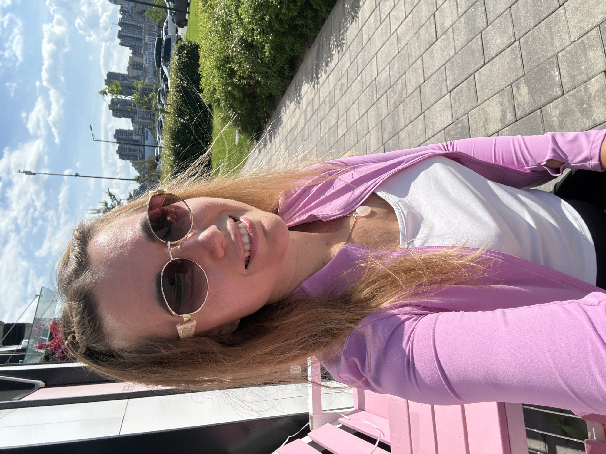 The image is a well-lit outdoor selfie of a young woman taken in an urban environment during daylight hours. The woman, with light brown to blonde hair, is smiling pleasantly at the camera. She is wearing round sunglasses with gold-colored frames, a white sleeveless top, and a light purple long-sleeved jacket or cardigan. A white necklace with a pale, possibly rose quartz, pendant hangs around her neck. The word "Dior" is faintly visible on the arm of her sunglasses.
She appears to be sitting or leaning against a distinct bright pink, slatted bench or railing, with a white string or light wire draped across it. The background reveals a paved area with large, rectangular grey blocks, lush green bushes, and several cars parked along a street. In the distance, a skyline of tall buildings suggests a city setting. The sky is bright with scattered white clouds, indicating sunny weather, likely midday or early afternoon. The overall impression is one of a cheerful moment in an urban park or outdoor cafe area.