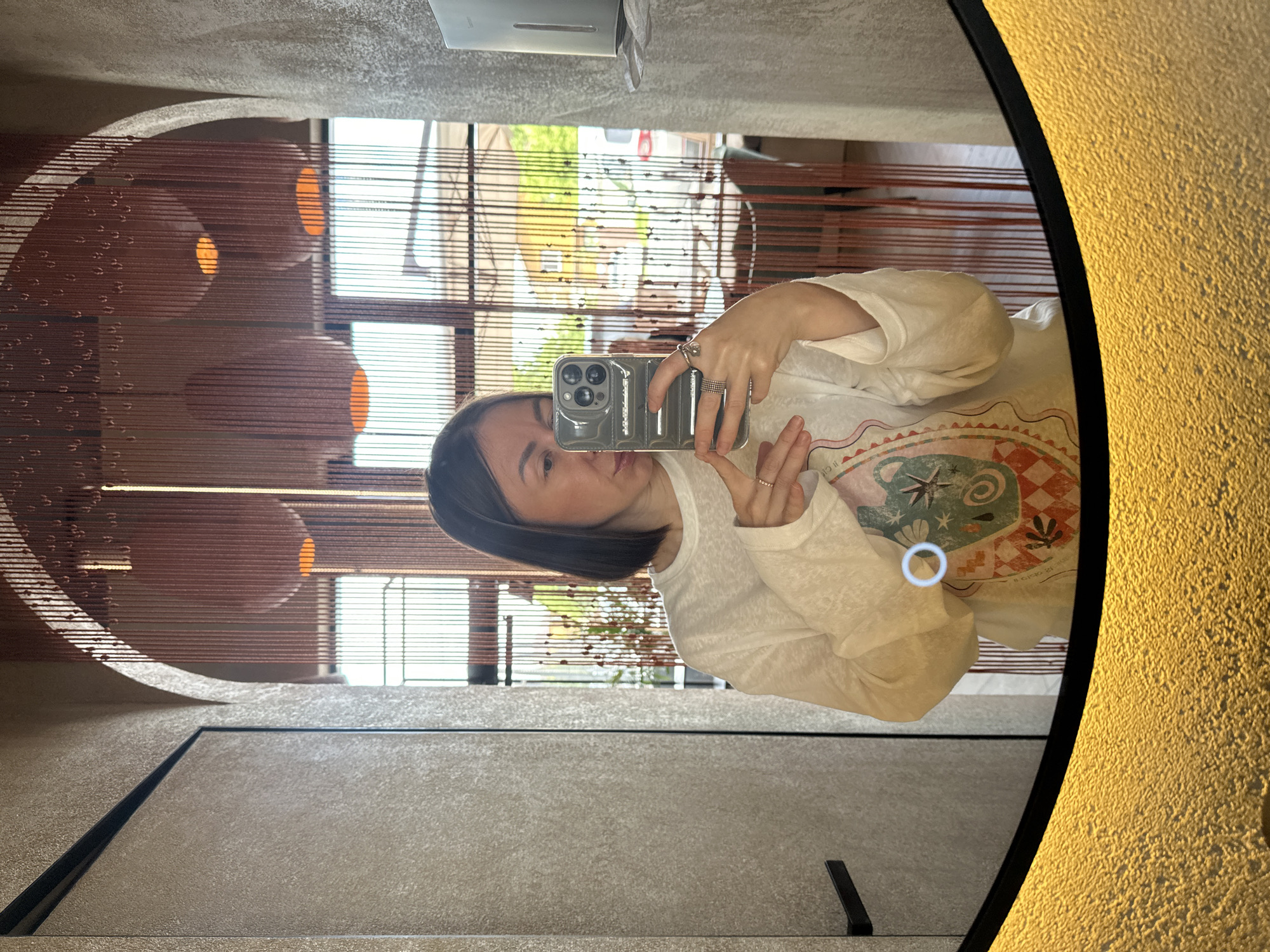 The image captures a mirror selfie taken by an adult female within a commercial establishment in Усады, Russia, likely a restaurant, cafe, or its associated restroom. The woman is positioned centrally, visible from the chest up, holding a dark gray smartphone vertically with both hands to capture her reflection. She has straight, dark, mid-length hair parted on her left side (viewer's right). Her facial features are partially obscured by the phone, but her right eye, nose, and mouth are discernible as she appears to be looking at the phone screen. She wears a white, long-sleeved top with a translucent quality, featuring a colorful graphic design on the lower left, depicting abstract patterns, a star, and checkered elements. On her left hand (viewer's right), she wears a prominent silver-toned ring with a central stone on her index finger, multiple stacked band rings on her middle finger, and a thin band ring on her ring finger. Her right hand (viewer's left) displays a single thin band ring on the ring finger. The smartphone, likely an iPhone, features a triple camera array on its rear and is housed in a textured, ribbed protective case. The mirror itself is oval or rounded-rectangular with a thick black frame along its bottom edge. This lower section of the mirror emits a warm, textured golden light. A small, circular, illuminated blue-white touch control or light source is reflected on the mirror's surface near the woman's lower left arm. The background, visible in the mirror's reflection, details the interior and exterior environment. The walls appear textured: a light brown/beige on the left with a distinct dark vertical line, possibly a door frame, and a black horizontal handle, suggesting a door. On the right, the wall is a lighter gray textured surface, featuring a wall-mounted silver-colored soap or sanitizer dispenser. An arched opening is visible in the upper right section, leading to another internal area. Directly behind the woman, and extending across the upper portion of the reflection, is a decorative bead curtain composed of numerous thin, reddish-brown strings with small, teardrop-shaped beads. Through and above this curtain, several large, spherical or oblong pendant lights are visible, emitting a warm orange glow from their bases. Beyond the bead curtain, a window or open area reveals an outdoor scene under bright daylight. Green foliage, an outdoor seating area with tables and chairs, and a partially visible white vehicle (possibly a
