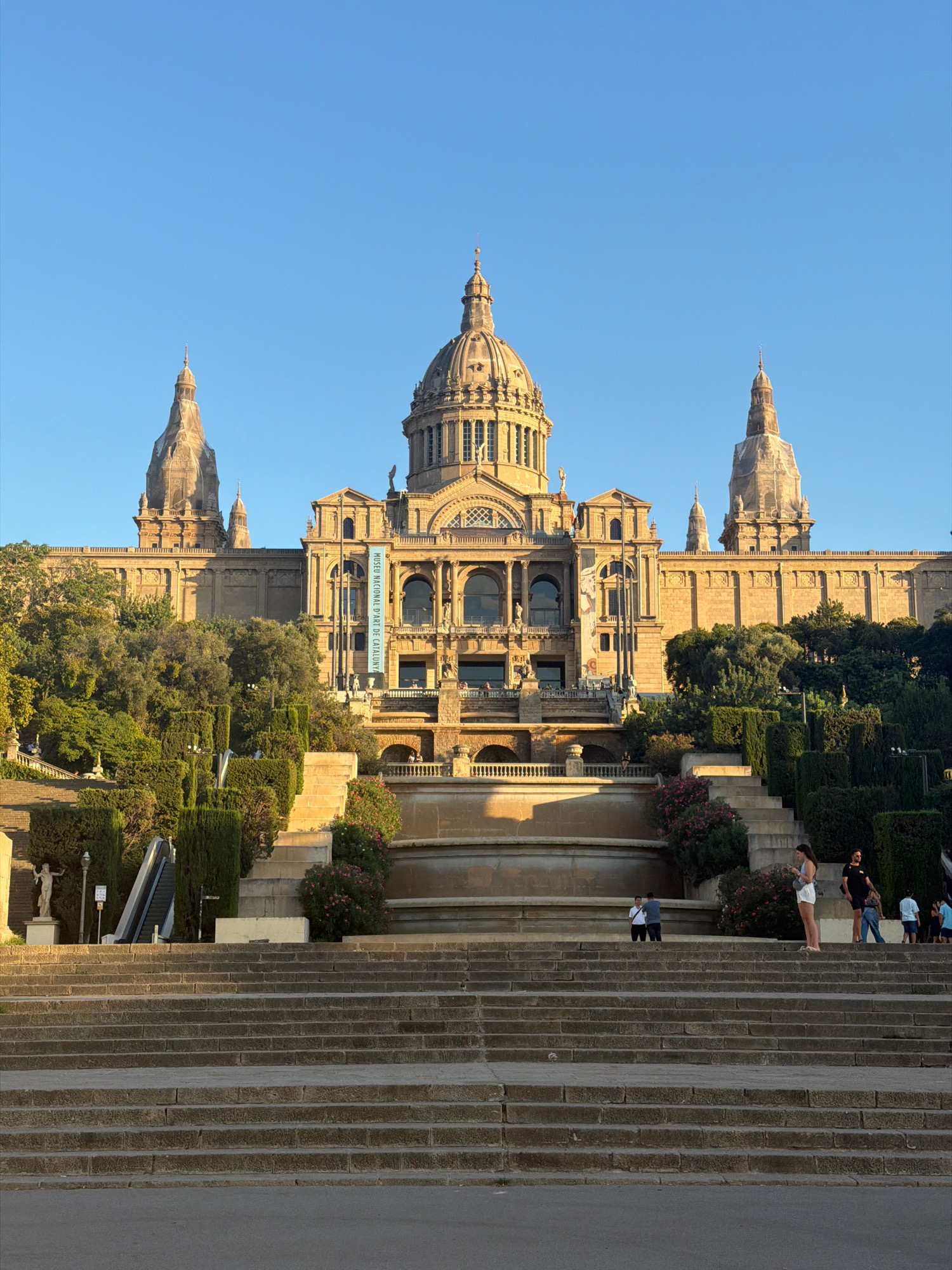 The image captures the iconic Palau Nacional, the National Palace of Catalonia, situated on Montjuïc hill in Barcelona, Spain. The grand building, with its central dome and flanking towers, is bathed in the warm light of the setting sun. In the foreground, a wide, stone staircase leads up towards the palace, with several people scattered across its steps. Some are walking, others are pausing to admire the view, and a few children are playing. Lush green trees and manicured hedges surround the staircase and cascade down the hillside, adding a touch of nature to the architectural splendor. A banner on the palace facade reads "MUSEU NACIONAL D'ART DE CATALUNYA". The sky above is a clear, vibrant blue, indicating a beautiful day.