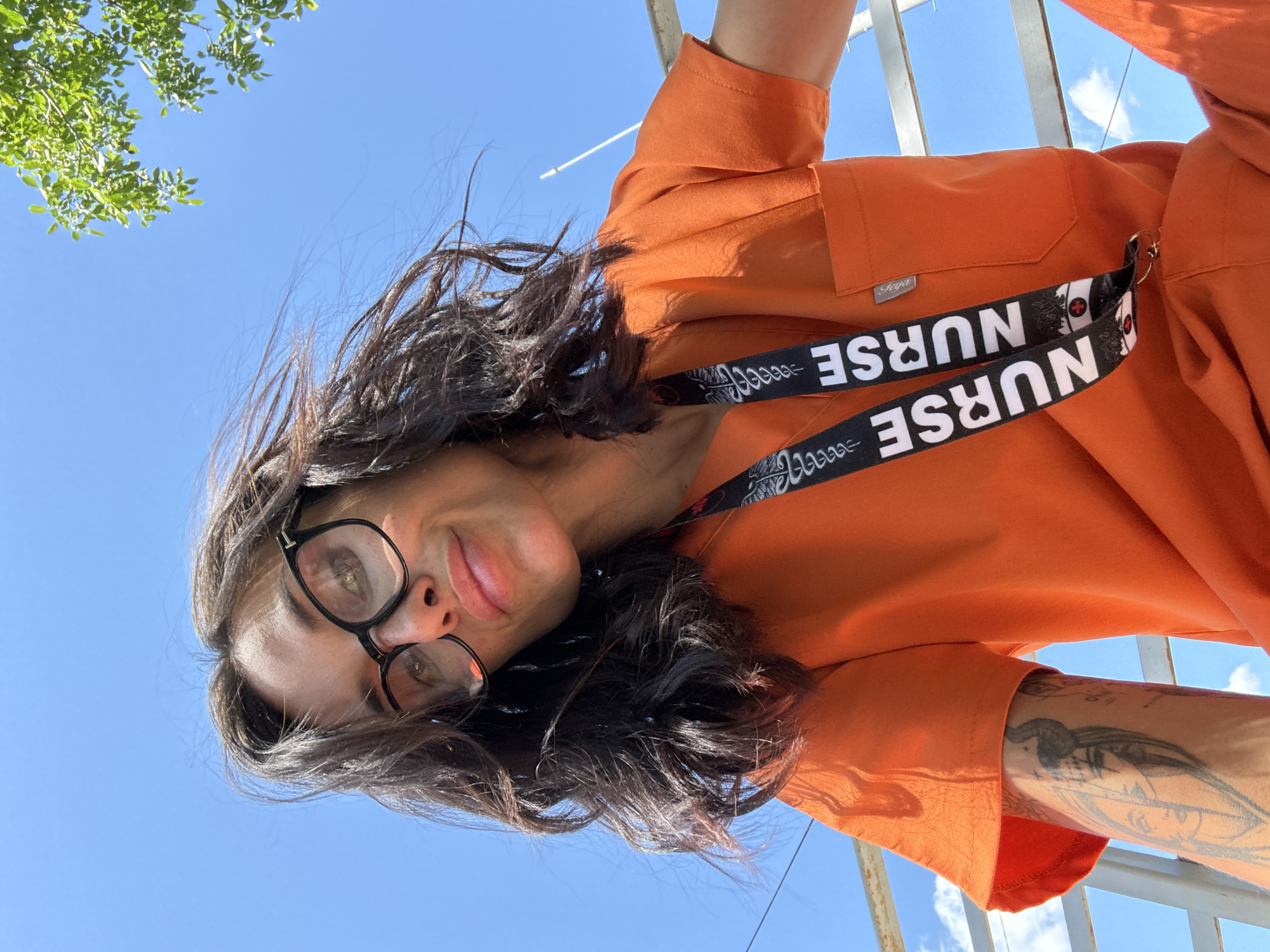 The image shows a low-angle, close-up selfie of a woman wearing black-rimmed glasses and an orange scrub top. Her dark, wavy hair is blowing in the wind. A lanyard with the word "NURSE" printed on it is visible around her neck. A tattoo of a woman with horns is visible on her left arm. The background consists of a bright blue sky with some white clouds and a glimpse of green leaves from a tree. The woman appears to be outdoors, possibly on a balcony or elevated area, with metal railings visible in the foreground. The scene is likely situated in Pleven, Bulgaria, given the context provided.