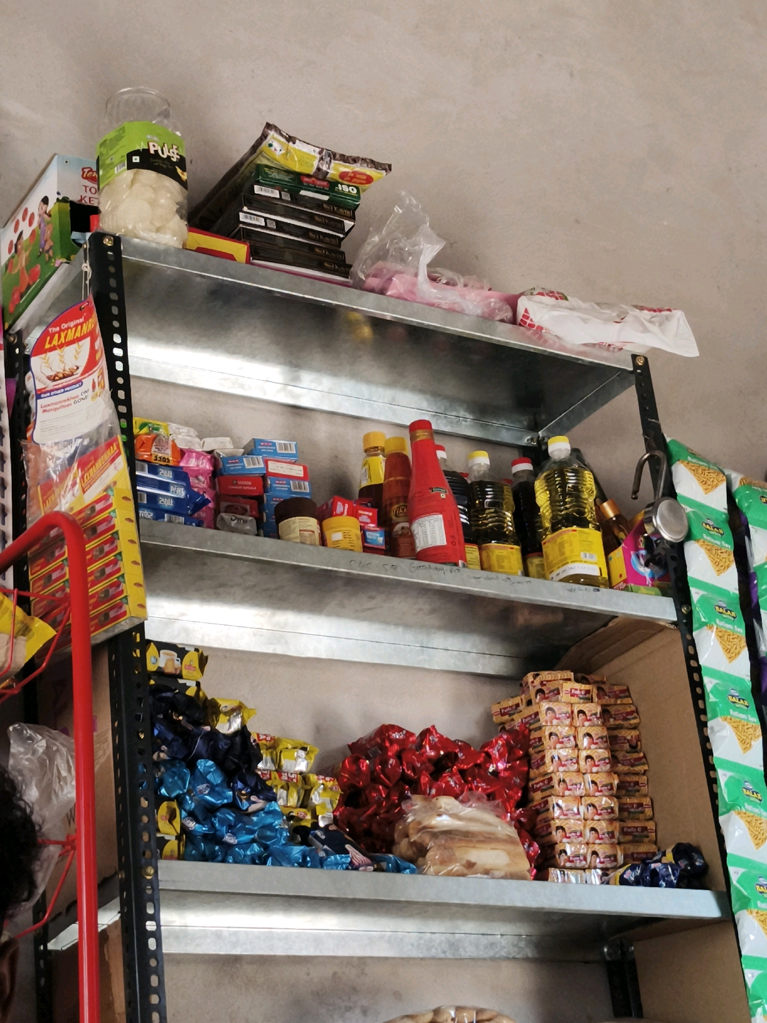 This is a medium-shot, eye-level photograph of a small convenience store shelf stocked with various grocery items. The scene is well-lit with artificial light, and the overall impression is one of organized clutter.
On the top shelf, a large clear plastic container filled with white puffed rice or snacks is prominent, labeled "Puls." To its right, a stack of black and green boxes are piled, and behind them, some crumpled plastic packaging.
The second shelf from the top holds a variety of packaged snacks and food items. Prominently displayed are several red and blue packets, likely snacks or biscuits. There are also smaller containers, possibly spices or condiments, and a few bottles of cooking oil, one of which is particularly large and yellow. A red bottle, likely ketchup, is also visible.
The third shelf is packed with more packaged goods, primarily in blue and yellow wrappers. A large pile of red wrapped items, possibly candy or snacks, dominates the center of this shelf. To the right, a stack of small, individually wrapped food items, possibly biscuits or snacks, are neatly arranged in rows. These packages feature images of people.
The bottom shelf, partially visible, contains more packaged snacks. On the far right, several green and white packets of snacks are hanging vertically, with prominent branding in green.
A dark gray metal shelving unit supports these shelves. A bright red metal rack is visible on the left side of the image. The background is a plain, light-colored wall, suggesting an indoor setting.
The image appears to be taken in Taranagar, India, as indicated by the context. No people are clearly visible in the frame, though the arm and shoulder of an individual are partially visible on the far left, and the back of another person's head is visible in the lower left corner. The scene depicts a typical retail environment for everyday consumer goods.