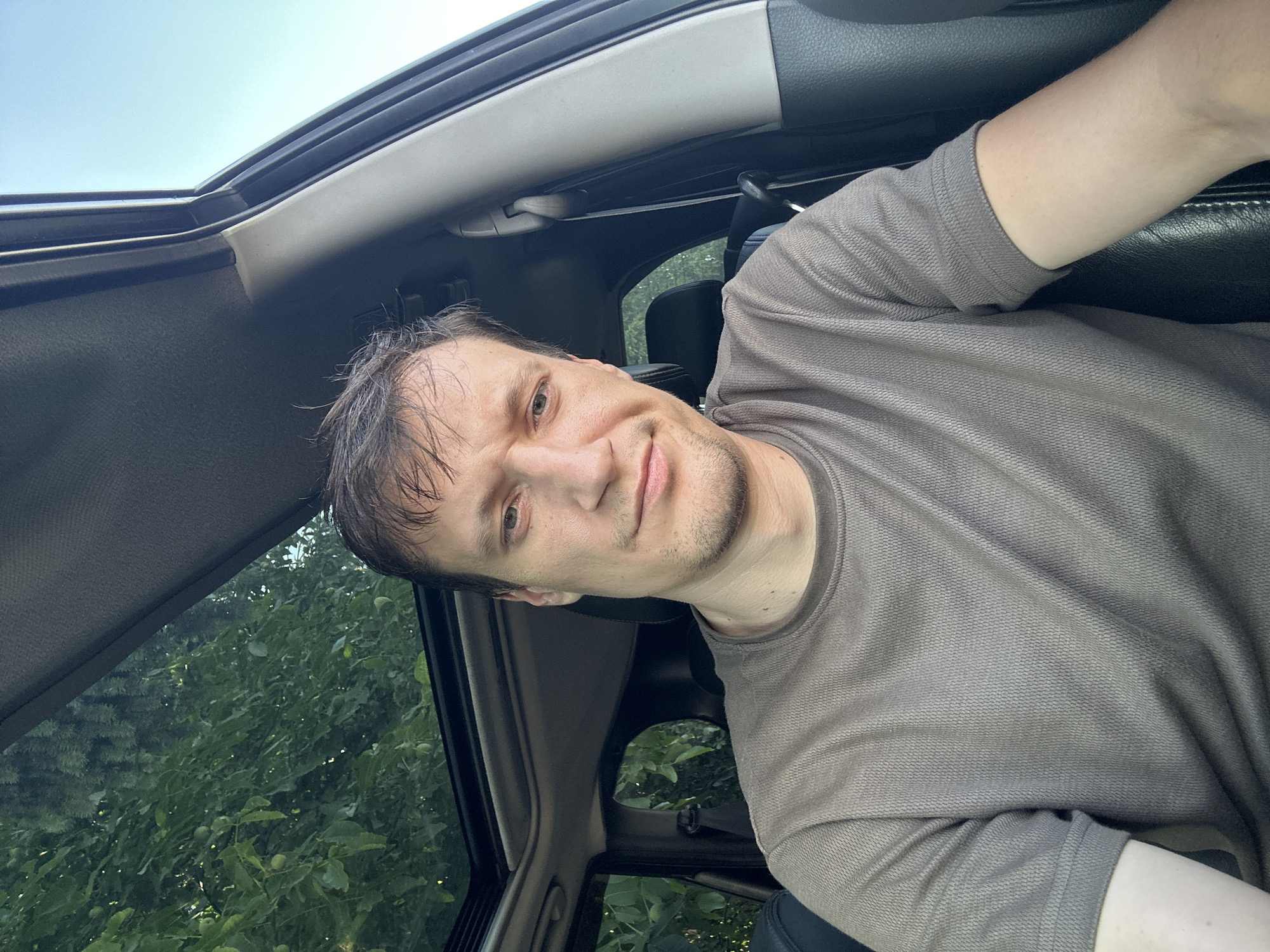 A man is sitting in the driver's seat of a car, looking at the camera. He has short, dark brown hair and is wearing a brown t-shirt. The car has a sunroof, which is open, and through it, green trees and a blue sky are visible. The interior of the car is dark, with black leather seats. The man appears to be in his late 20s or early 30s. The image was taken outdoors, with natural lighting. The man's facial expression is neutral. The location is not specified in Shatava, Ukraine.