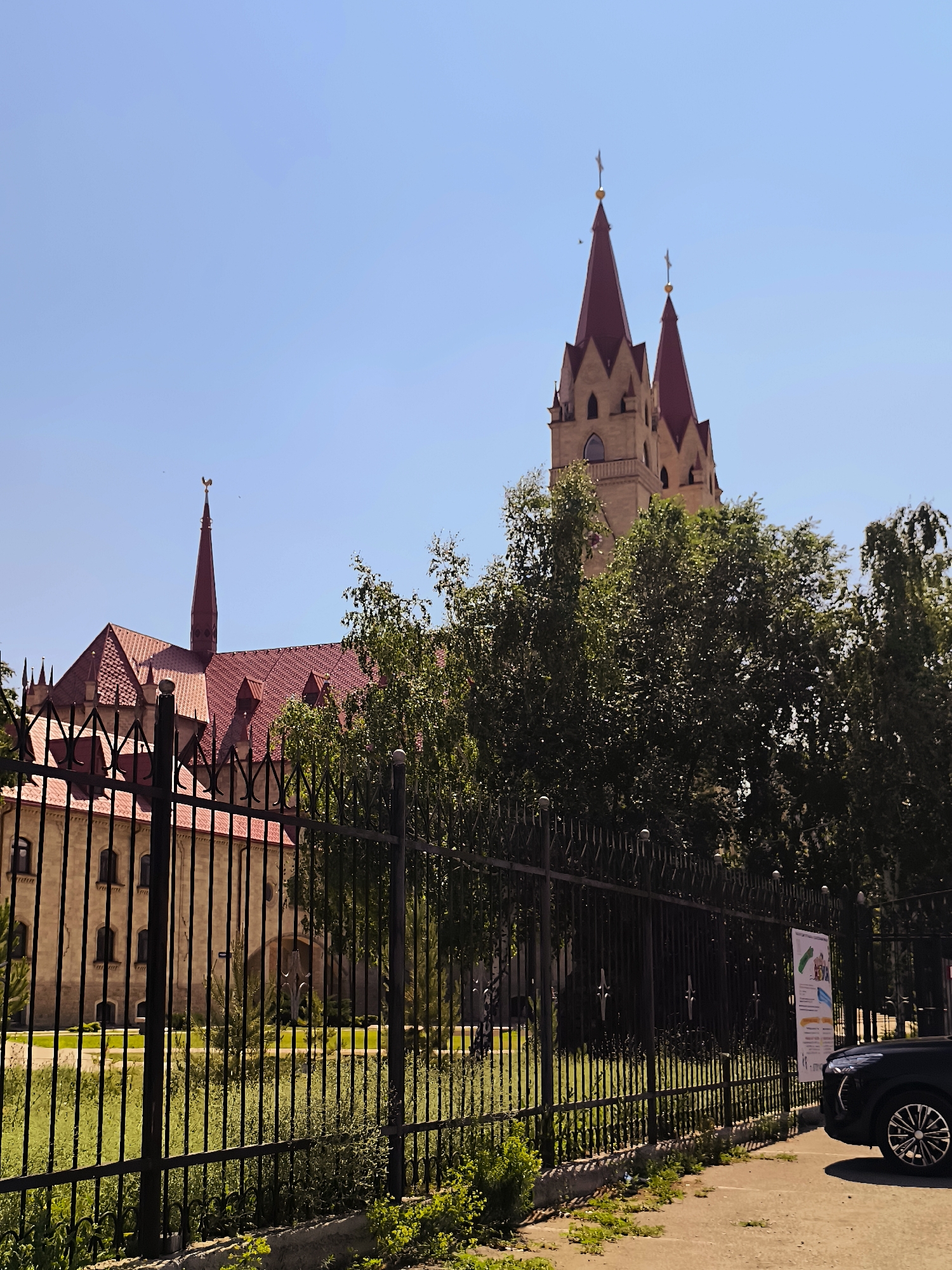 The image depicts a segment of the Qarağandı City Administration building in Kazakhstan, specifically its distinctive architectural features. The building exhibits a neo-Gothic style with a prominent red-tiled roof. Several spires adorn the structure, two of which are particularly tall and topped with crosses. A smaller spire, featuring a golden rooster weather vane, is visible on the left side. The building's exterior is constructed of light-colored stone, with arched windows. A black metal fence runs across the foreground, separating the viewer from the building and a patch of green grass. Trees with lush green foliage partially obscure the lower levels of the administration building. In the lower right corner, the front quarter of a black modern car is visible, parked on a paved surface. A poster is attached to the fence to the right of the car. The sky is a clear, light blue, indicating a sunny day.