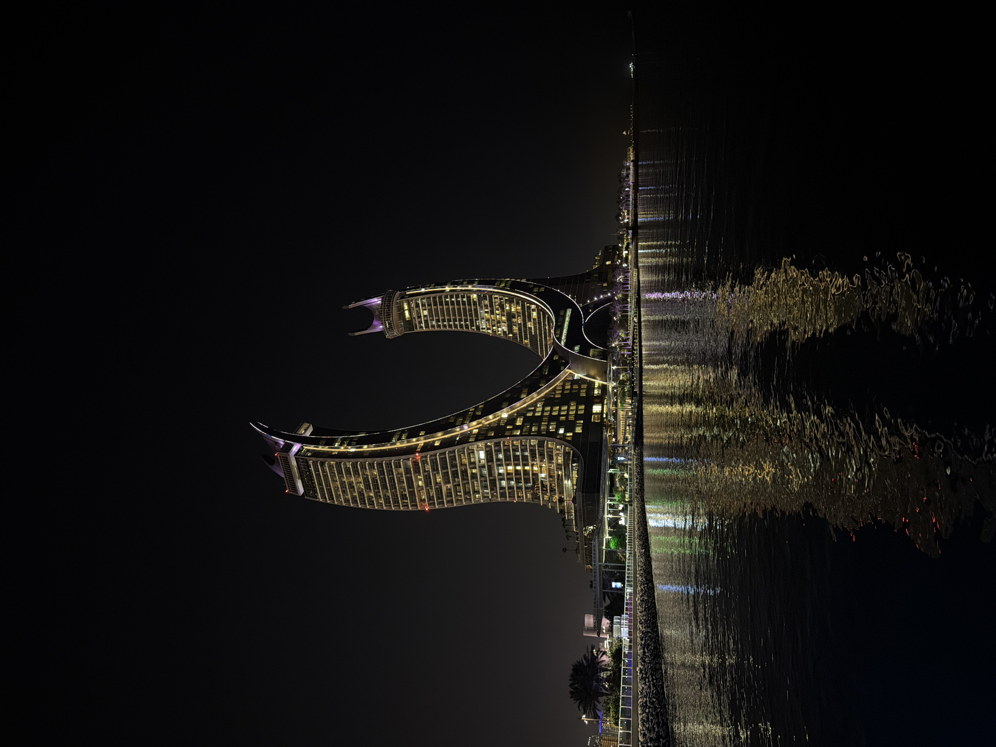 This is a nighttime, vertical, eye-level photograph of the iconic Tornado Tower in Doha, Qatar, reflected in the calm waters of the Arabian Gulf. The skyscraper, illuminated by numerous lights, stands prominently against the dark sky, its distinctive curved structure creating a dramatic silhouette. The building's exterior is a mix of glass and metal, with warm yellow lights emanating from within, highlighting its architectural details. The water in the foreground acts as a mirror, reflecting the vibrant lights of the tower and the surrounding cityscape, creating a dazzling display of color and light. The reflection is somewhat distorted by the gentle ripples on the water's surface, adding a dynamic element to the otherwise still scene. A stone embankment with a white railing lines the edge of the water, with palm trees and other vegetation visible in the distance. The overall impression is one of modern elegance and serene beauty, showcasing the architectural marvel of the Tornado Tower at night.