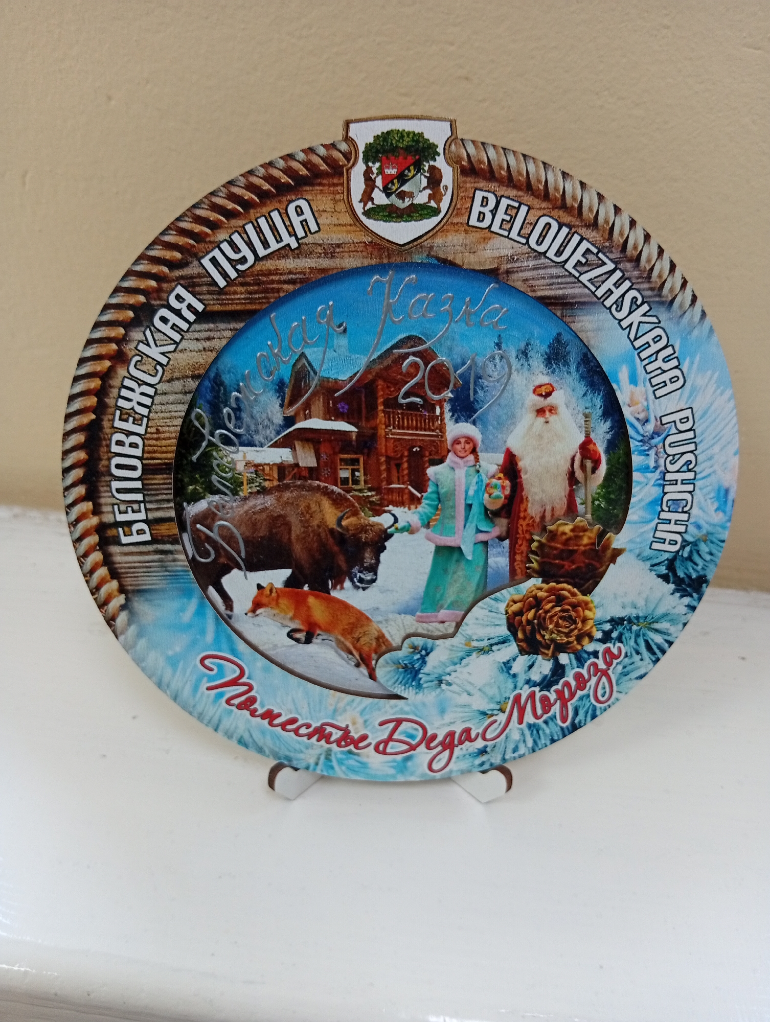 The image shows a circular wooden plaque with a festive winter scene. The plaque features a rope-like border. In the center, a stylized depiction of Belovezhskaya Pushcha National Park in Belarus is presented. The artwork includes a wooden house, snow-covered trees, a European bison, a fox, Santa Claus (Ded Moroz) in traditional red and white attire, and a young woman in a blue winter coat and hat, possibly representing Snegurochka. The year "2019" is prominently displayed. Text in Russian surrounds the central image, with phrases like "Пуща", "Беловежская", and "Деда Мороза". There are also two pine cones depicted on the plaque. The plaque is propped up by two small white stands. The background is a plain, light-colored surface, possibly a shelf or table, and a wall.