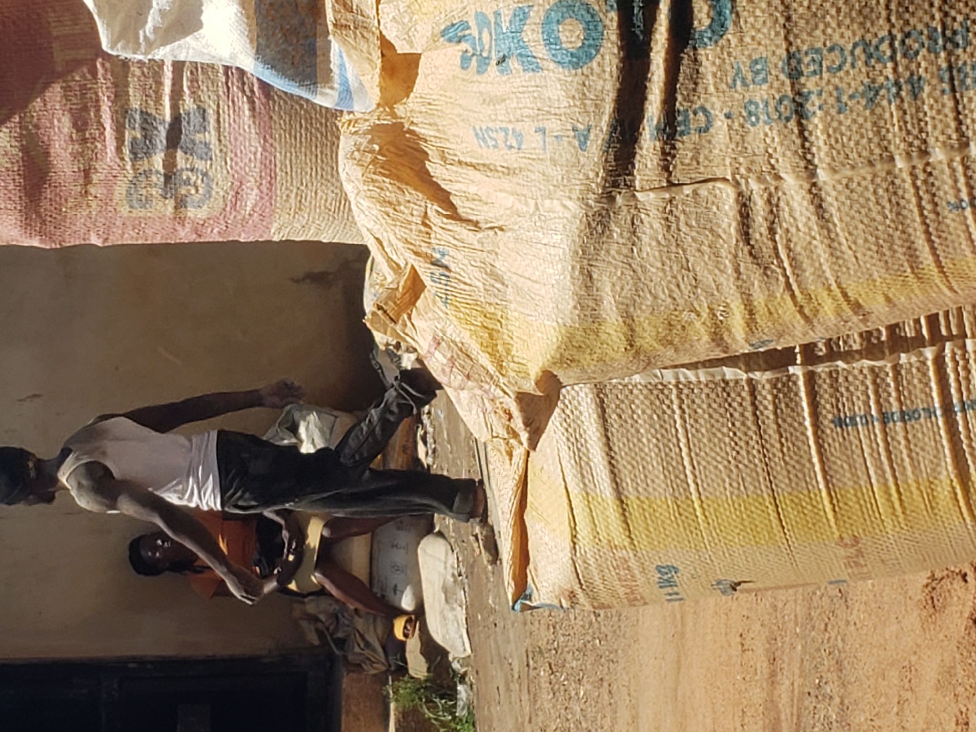 The image depicts an outdoor scene, likely in Jos, Nigeria, with a focus on large sacks of what appears to be cement or grain, common in commercial areas. In the background, a woman in an orange top and yellow skirt is seated on a sack, while a man, wearing a white tank top and dark pants, walks away from her. Both individuals have dusty skin, suggesting an environment where dust is prevalent. The foreground is dominated by several large, textured sacks. One prominent sack in the foreground is a beige color with yellow markings and text, including "10 KG". Another sack, partially visible behind it, is red and yellow with blue lettering that appears to be "B...CO." The ground is dusty and appears to be unpaved. The overall impression is of a storage or trading area in an arid or dusty urban environment.
