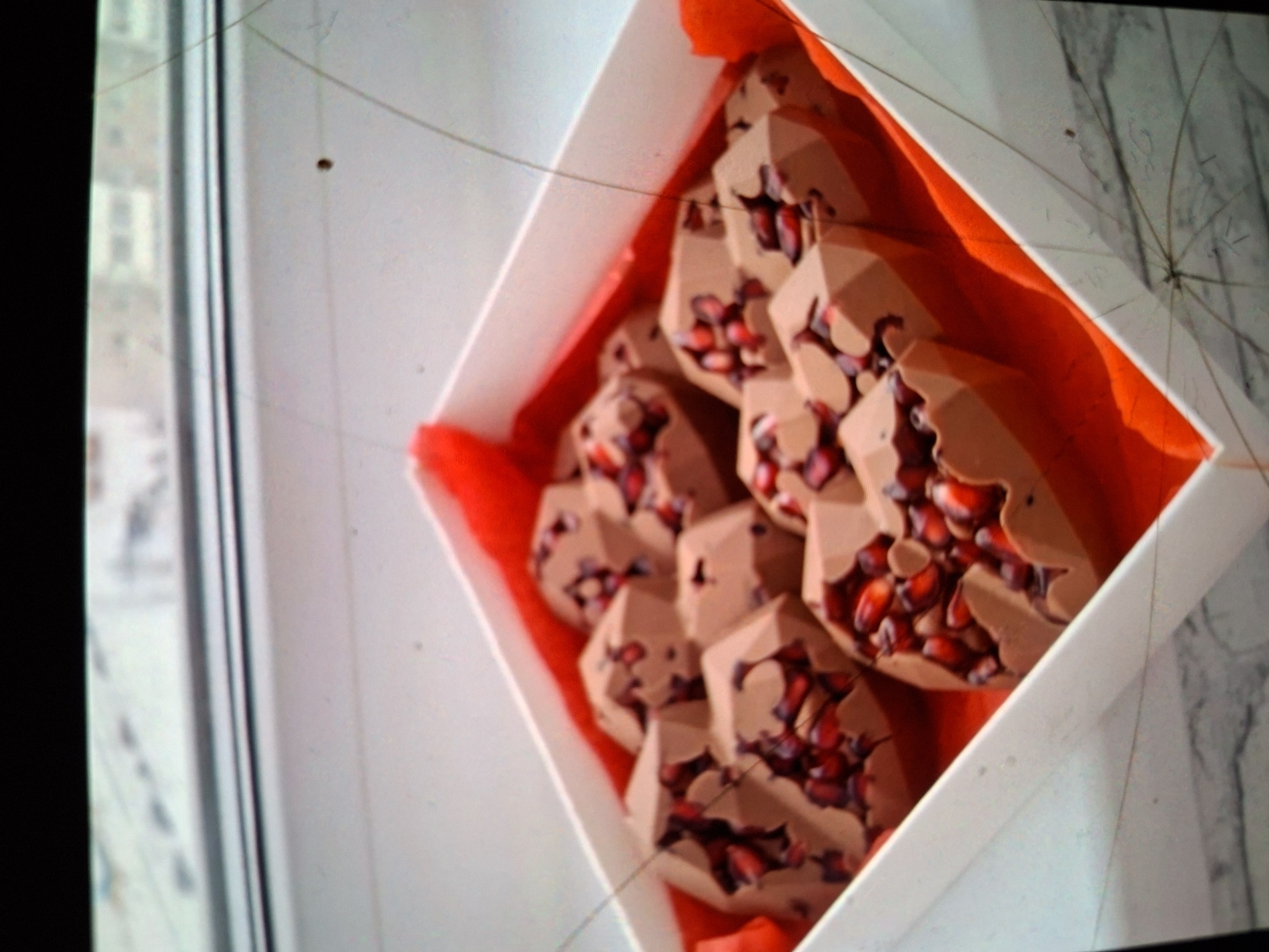 A white box filled with faceted chocolate candies, each embedded with pomegranate seeds, is positioned on a windowsill. The box is lined with bright red fabric. Outside the window, a blurred cityscape of buildings is visible. The scene appears to be indoors, likely a domestic setting, in the city of Medvezhyi Ozyora, Russia.