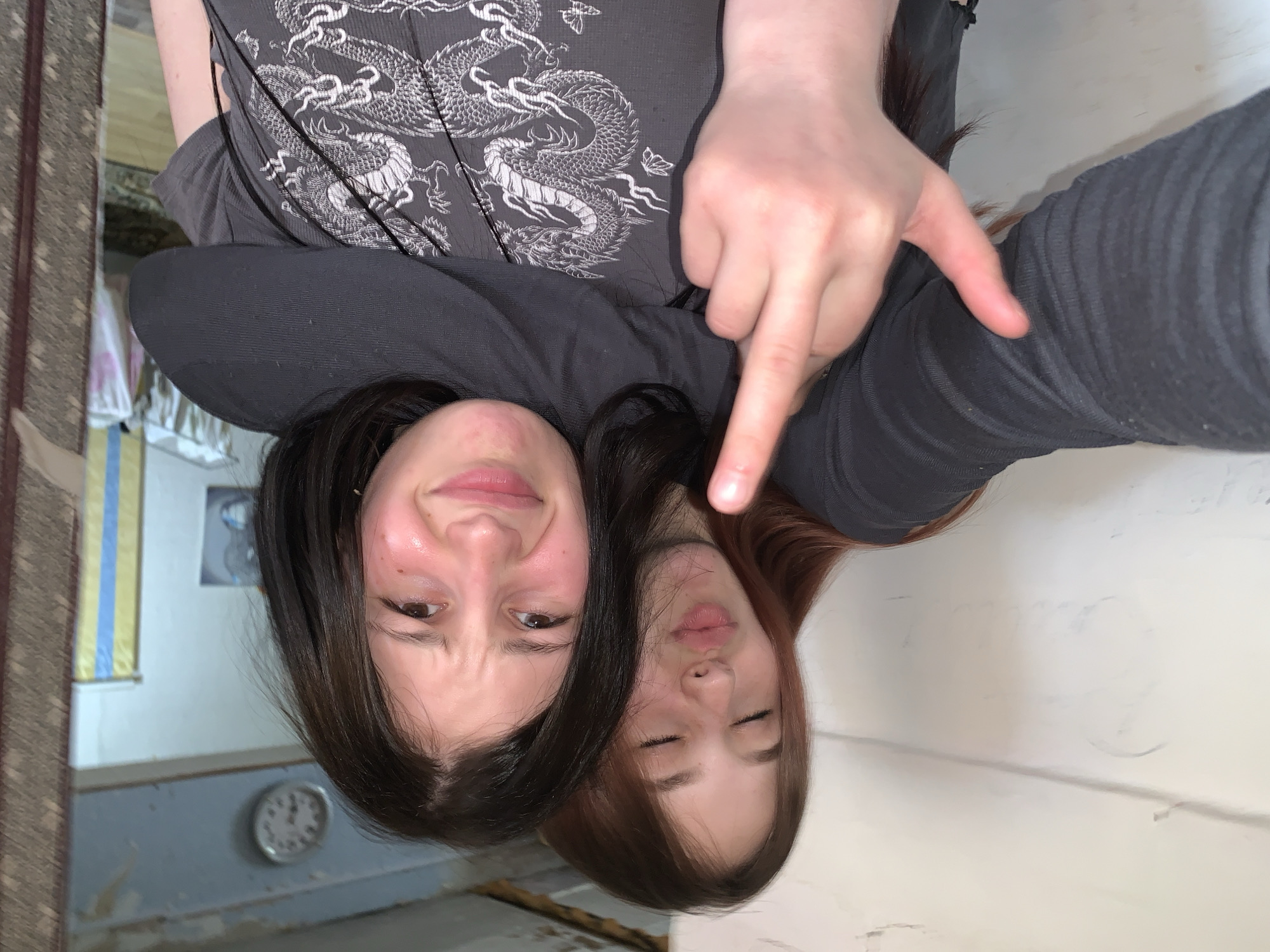An informal indoor selfie captures three people, likely friends, in a casual setting in Фоки, Russia. The image is taken from a top-down perspective, looking down at two young women. One woman, with dark hair, is positioned on the left and has a playful expression with a slight smirk. Her cheeks appear flushed. Next to her, on the right, is another young woman with reddish-brown hair, eyes closed and lips slightly parted, appearing relaxed or asleep. A third person, likely the one holding the camera, is partially visible at the top, wearing a dark grey t-shirt adorned with a detailed white dragon and butterfly design. Their right hand is prominent, pointing downwards towards the two women's faces.
The background on the left reveals a domestic environment, featuring a wall-mounted analog clock showing approximately 11:55 or 12:00. Above the clock, a section of wall with vertical yellow and blue stripes is visible, possibly part of a curtain or wallpaper. Further left, a patterned rug or carpet can be seen. The right side of the background is a plain white wall. The overall lighting suggests an indoor setting, possibly during the daytime. The interaction appears friendly and intimate, capturing a candid moment between the individuals. There is no visible text in the image.