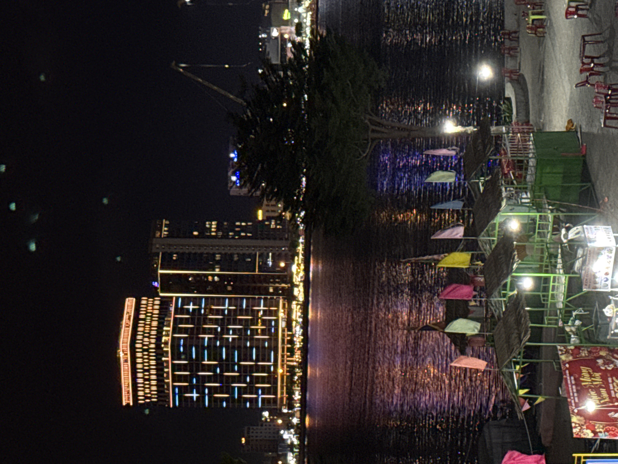 This is a nighttime scene on the Han River in Da Nang, Vietnam, looking across the water towards the city skyline. In the foreground, there are several small, open-air stalls with thatched roofs, some illuminated by green lights. Colorful flags, including pink, yellow, light blue, and white, fly above the stalls. A large banner with Vietnamese text, possibly a New Year greeting, is visible on the left. The river reflects the lights of the city, creating a shimmering effect. Across the river, a modern, illuminated skyscraper with a grid-like pattern of lights dominates the skyline, flanked by other buildings and a construction crane. The night sky is dark with some faint green lights, possibly bokeh from the camera lens. The overall atmosphere is vibrant and festive, suggesting a lively waterfront area.