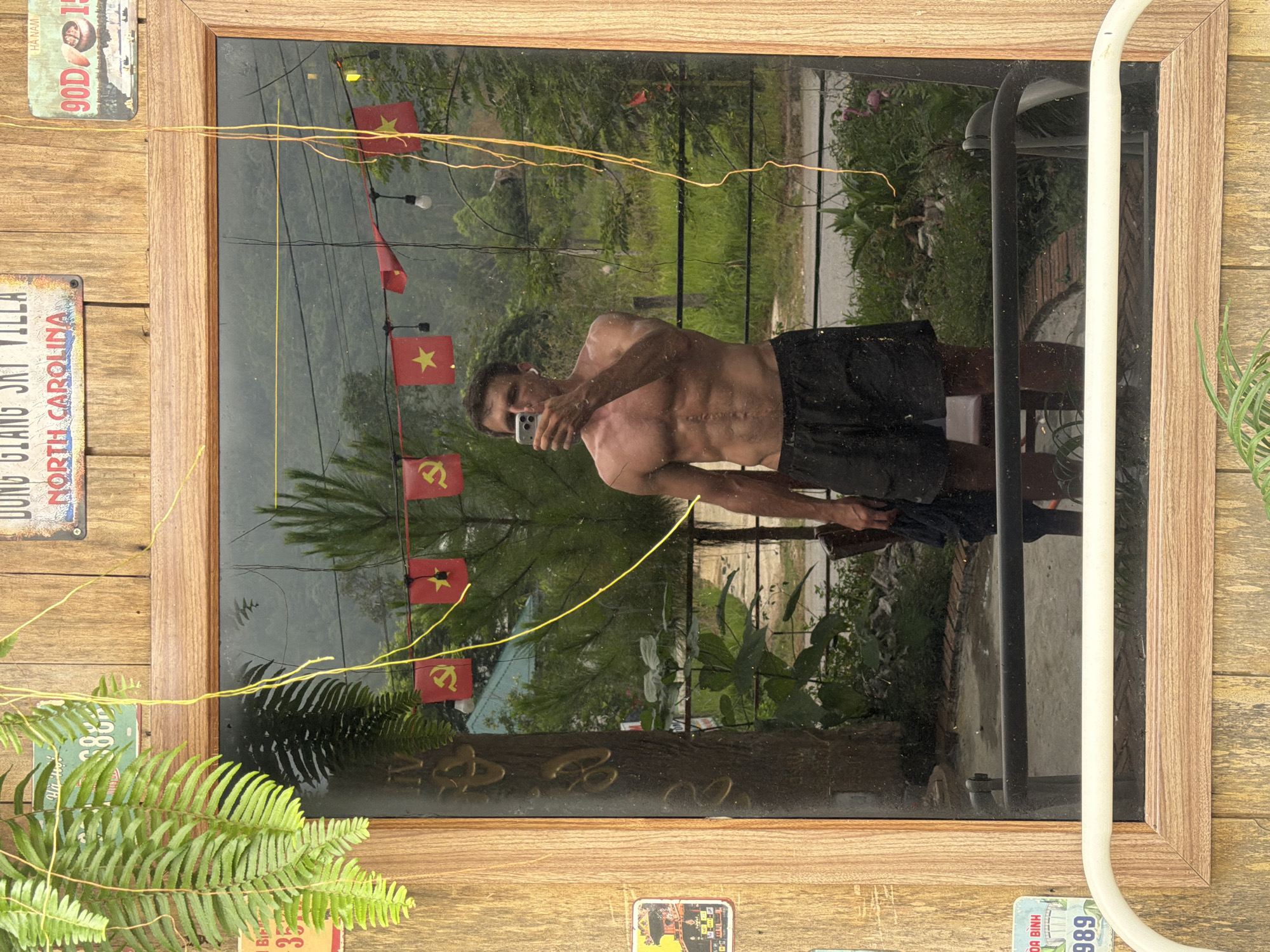 The image shows a mirror framed in wood, reflecting a man taking a selfie. The man is shirtless and wearing dark shorts. His reflection also reveals Vietnamese flags hanging in the background. The mirror is mounted on a wooden wall decorated with various license plates and greenery. The reflection in the mirror includes a glimpse of a street scene and lush vegetation, characteristic of Đà Nẵng, Vietnam. A white, curved metal structure is adjacent to the mirror's right side.