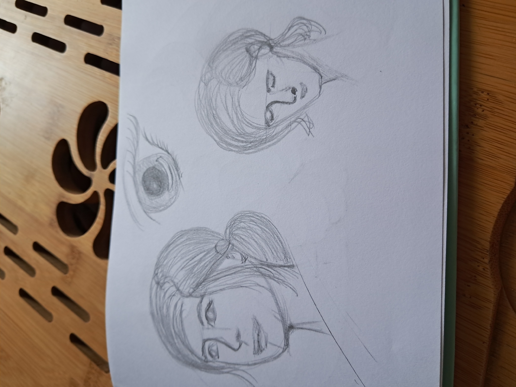 A pencil sketch of three portraits of women, rendered on white paper, rests on a wooden surface. The sketch features two full faces and an eye. The woman on the left is facing left and appears to be smiling. Her hair is pulled back in a bun. The woman on the right is looking down with her eyes closed, her hair also styled back. The eye is drawn separately above and to the right of the two faces. The wooden surface has a slatted design with carved floral patterns.