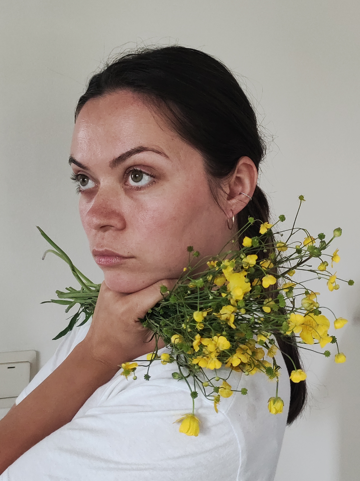 A close-up portrait captures a woman with dark hair pulled back, positioned against a plain, light-colored wall, likely indoors in Фоки, Russia. She is depicted from the chest up, wearing a simple white T-shirt. Her left hand is raised to her chin, cradling a bouquet of vibrant yellow wildflowers, possibly buttercups, with long green stems and leaves. The flowers are casually arranged, draping over her hand and slightly onto her shoulder.
The woman has fair skin with visible texture and light freckles across her nose and cheeks. Her dark eyebrows frame expressive green eyes, which are cast upwards and to her left, conveying a contemplative, pensive, or somewhat solemn expression. She wears two small silver hoop earrings and a twisted silver ear cuff in her visible left ear.
The background is stark and uncluttered, with a small white electrical fixture partially visible on the lower left side, behind her arm. The lighting is soft and even, suggesting either natural light from an unseen window or diffused indoor lighting. The overall mood is quiet and introspective. No text is visible in the image.