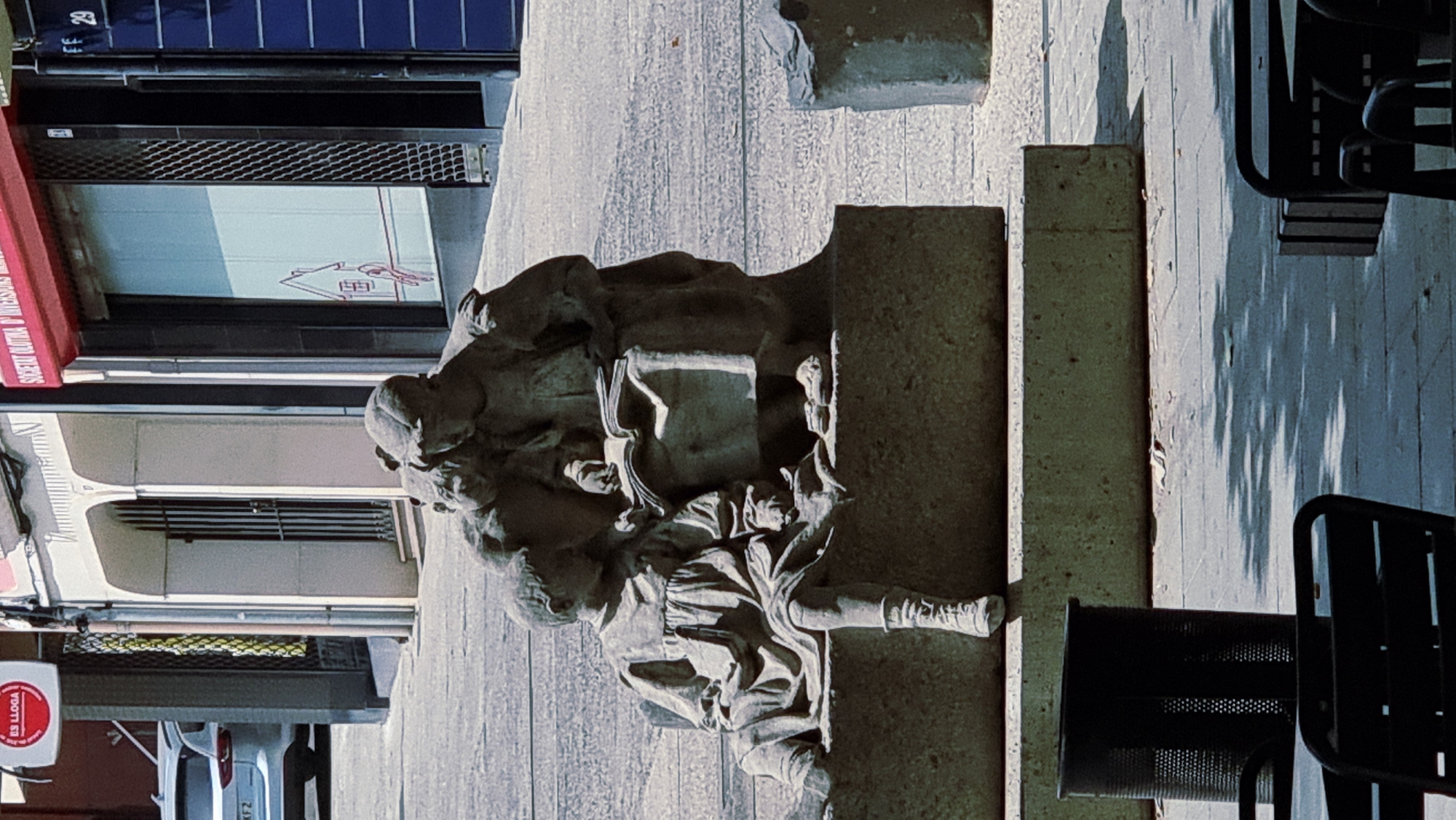 The image shows a bronze statue of three children seated on a stone bench. The child on the left, a girl, is facing left and looking down. She is wearing a long dress and high-laced boots. The other two children are on the right, with one child holding an open book and pointing at it, while the third child leans in to look. The statue is located outdoors in a city setting. In the background, there are buildings with storefronts and a white car parked on the street. A black trash can is visible to the left of the statue, and parts of dark outdoor furniture are in the foreground. Based on the presence of the "ES LLOA" sign, a business located in Olot, Spain, it is likely that this scene is in Olot, Spain. The overall lighting suggests it is daytime, with shadows cast by trees on the ground.