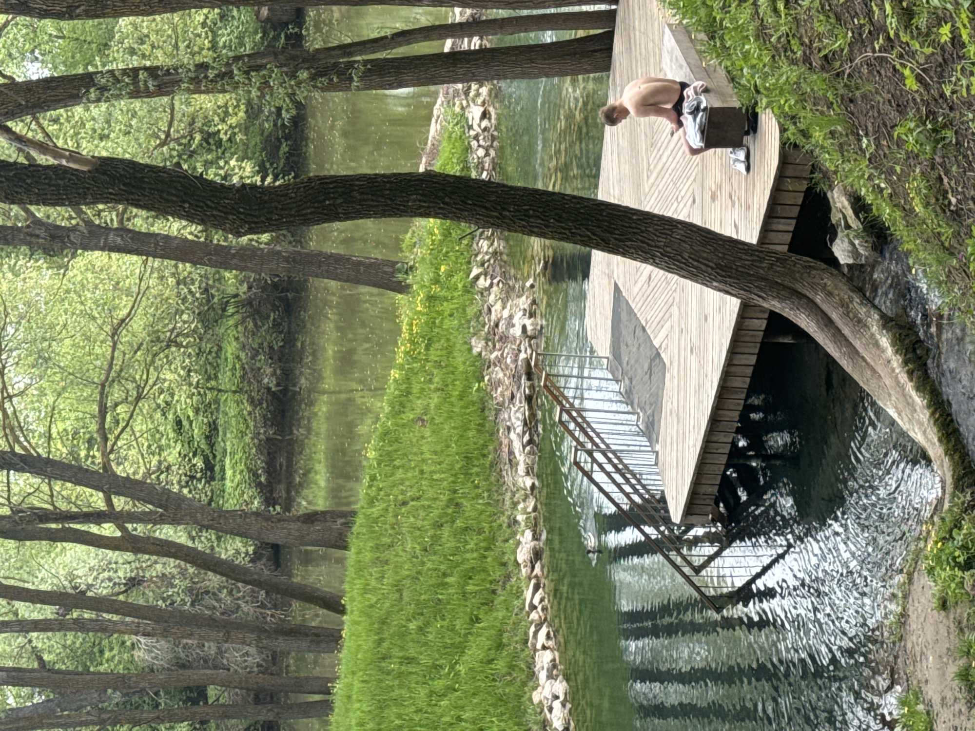A shirtless man sits on a wooden dock over a small river in Sdt Shcherbakovka, Russia. The dock has a metal staircase leading into the water. Trees line the riverbank. Grass grows on the edge of the water.