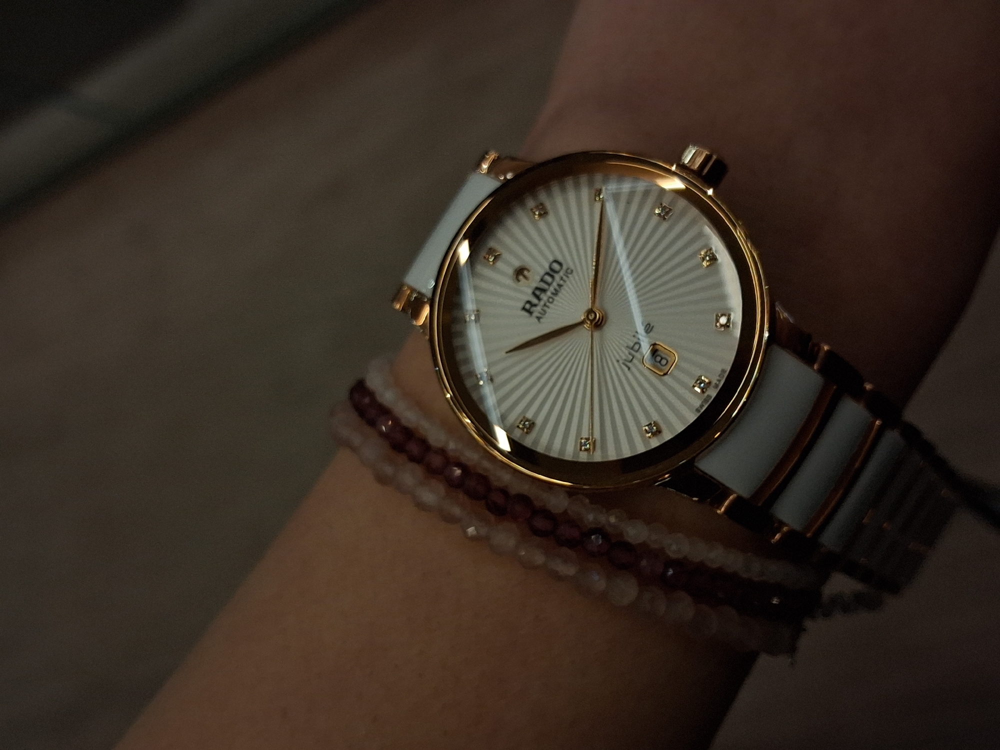 A close-up, downward-angled image captures a human wrist adorned with a Rado Automatic Jubile watch and two beaded bracelets. The watch features a white ceramic and rose gold-tone strap, a rose gold-tone bezel, and a white dial with a radial sunburst pattern emanating from the center. Diamond hour markers are set at the 1, 2, 4, 5, 7, 8, 10, and 11 o'clock positions, with the 3, 6, 9, and 12 o'clock positions marked by larger diamond clusters. A date window at the 6 o'clock position displays the numeral '8'. The watch hands are rose gold-tone. Positioned above the watch are two multi-strand bracelets: one composed of small, translucent, light-colored beads, and the other of darker, reddish-brown beads. The skin tone of the wrist is light to medium brown. The background is dark, out-of-focus, and lacks any discernible features. No specific location within Varna, Bulgaria, is identifiable from the visual information presented.