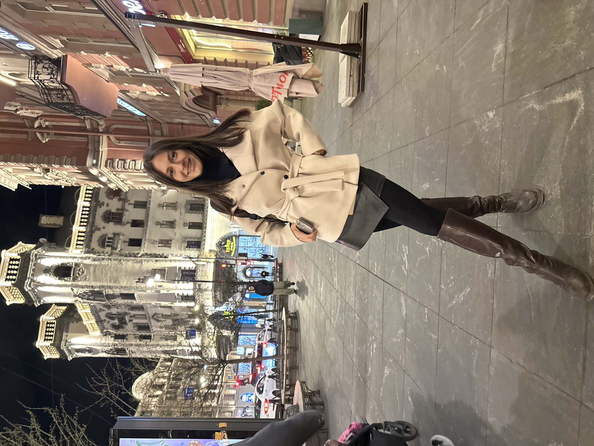 A young woman with long, dark hair stands smiling on a tiled city sidewalk at night, posing for a photograph. She is dressed in a light beige, belted trench coat or overcoat, a dark turtleneck, black leggings, and dark brown knee-high boots. She holds a small, light-colored object, possibly a phone, in her right hand.
The background showcases an illuminated urban setting with impressive multi-story buildings featuring ornate architectural details, including balconies, columns, and decorative facades, consistent with a historical European city. To the woman's left, a large, intricately lit building dominates the scene. Further down the street, various streetlights, cars with illuminated headlights and taillights, and other pedestrians indicate a lively evening environment. Trees with bare branches are visible, suggesting a cooler season.
On the paved sidewalk in the mid-ground, several circular tables and chairs, typical of outdoor cafe seating, are arranged. A red sign with white lettering displaying "TEATP" (Russian for "Theatre") is visible above a building in the background. The scene depicts a bustling street in Saint Petersburg, Russia, during the evening or night, under artificial light.