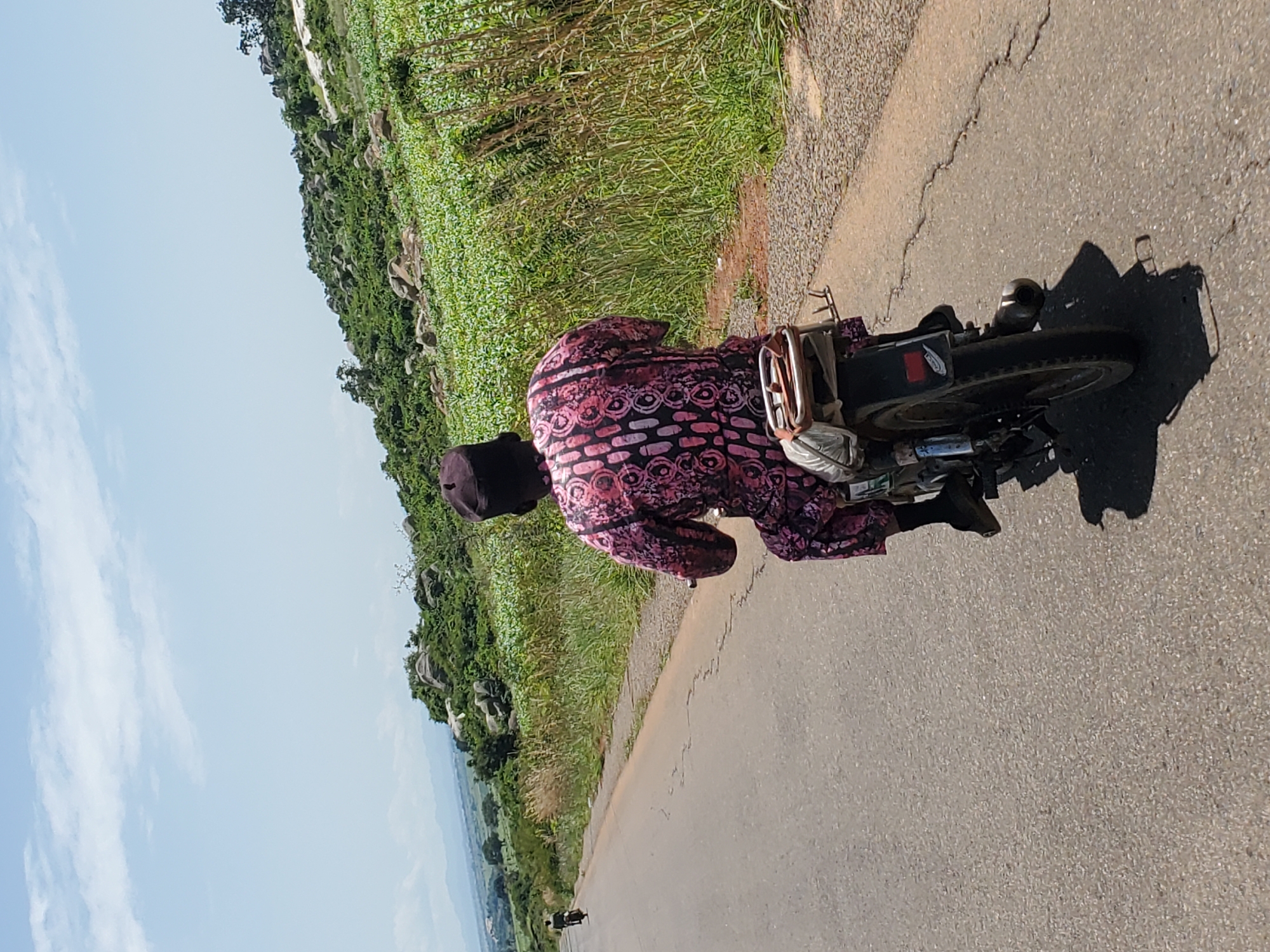 A person wearing a dark cap and a pink and black patterned outfit rides a motorcycle on an asphalt road. The motorcycle has a passenger seat with some bags attached. The road is bordered by tall green grass and a field of green crops on the right. In the background, there is a rocky, vegetated hillside. In the distance, mountains are visible under a partly cloudy blue sky. Another person on a motorcycle is further down the road to the left. The scene appears to be in a rural or semi-rural area, consistent with locations around Pankshin, Nigeria.