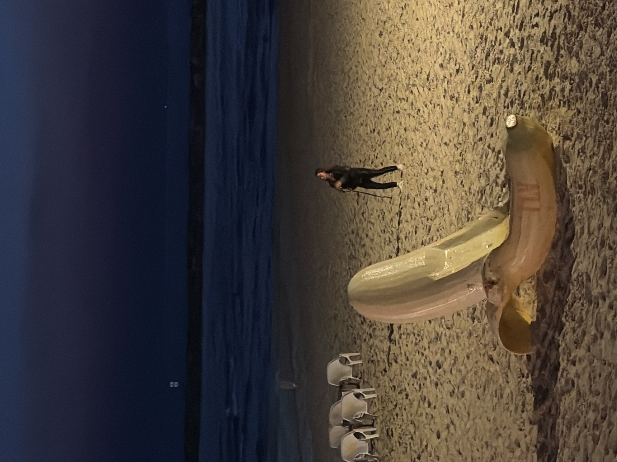A large, stylized sculpture resembling an open banana peel rests on a sandy beach. The sculpture is predominantly light yellow and pinkish-orange, with a textured surface. In the background, a person stands on the sand, holding a cane and looking upwards. The person is wearing dark clothing, including a jacket and pants, and white sneakers. Several empty, white beach chairs are arranged on the sand further back. The scene is illuminated by artificial lights, casting shadows on the sand. Beyond the beach, the ocean is visible with gentle waves, and the sky is dark blue, indicating it is either dusk or night. The location is the Tel-Aviv beach in Israel.