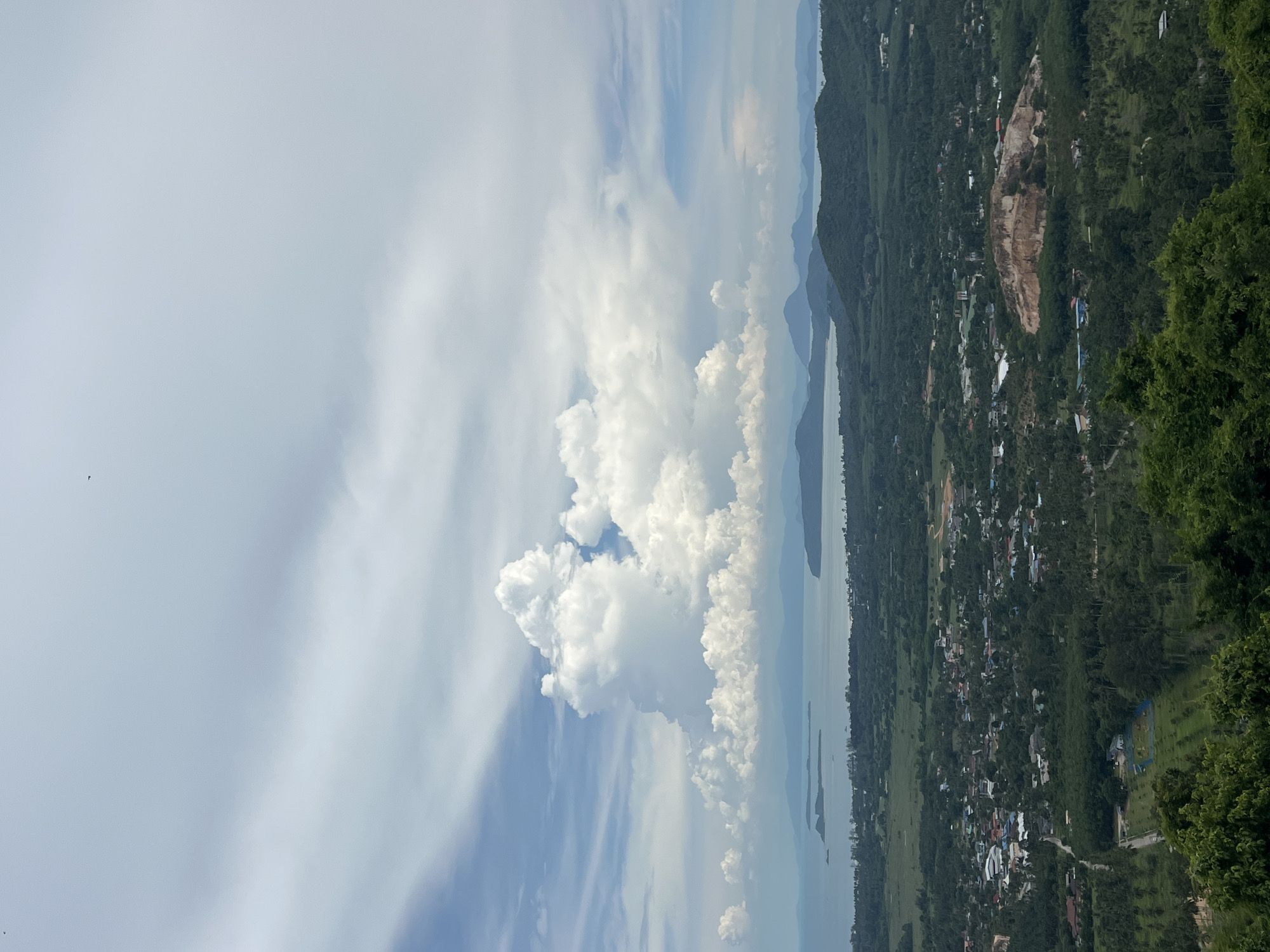An elevated, wide-angle, vertical shot captures a panoramic view of Baan Thurian, Thailand, under a dynamic sky filled with large, white cumulus clouds. In the foreground, the tops of lush green trees are visible. The midground reveals a sprawling landscape dominated by a small city nestled amongst dense, green vegetation. Buildings with a variety of colored roofs, roads, and what appear to be agricultural fields are scattered throughout. A prominent, lighter-colored hill or quarry site is visible in the middle ground, featuring exposed rock. Beyond the city and the varied terrain, a body of water, likely the sea, stretches to the horizon, with islands and distant, hazy mountains forming the backdrop. The sky is a mixture of blue and white, with the large cloud formation drawing significant attention due to its size and dramatic appearance. Two small, dark specks, possibly birds, are present in the upper portion of the sky. The overall impression is of a vast, natural landscape punctuated by human settlement.