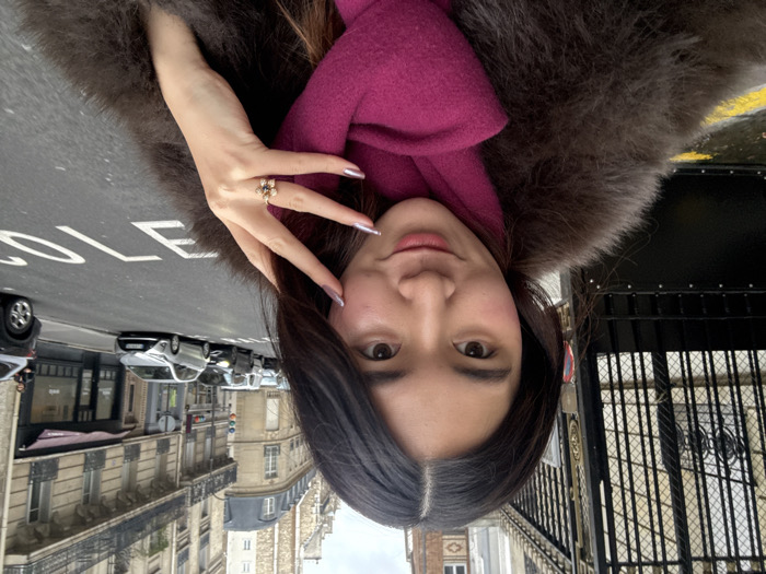 A medium-close-up selfie depicts a young woman standing on a city street in Paris, France. The woman, with long dark hair and a light complexion, looks directly at the camera with a subtle smile. She wears a dark brown faux fur coat and a bright magenta scarf. Her right hand is raised near her face, revealing metallic silver nail polish on her fingers and a flower-shaped ring on her ring finger.
In the background to her left, a large, ornate black wrought-iron gate with gold accents is partially visible. To her right, a paved street recedes into the distance, showing a line of parked and moving vehicles, including a prominent silver hatchback. The white word "ÉCOLE" (school) is painted in reverse on the asphalt. The street is flanked by classic Parisian Haussmannian buildings with light-colored stone facades, decorative balconies, and dark roofs. The sky is overcast, and a wet patch is visible on the ground near the gate.