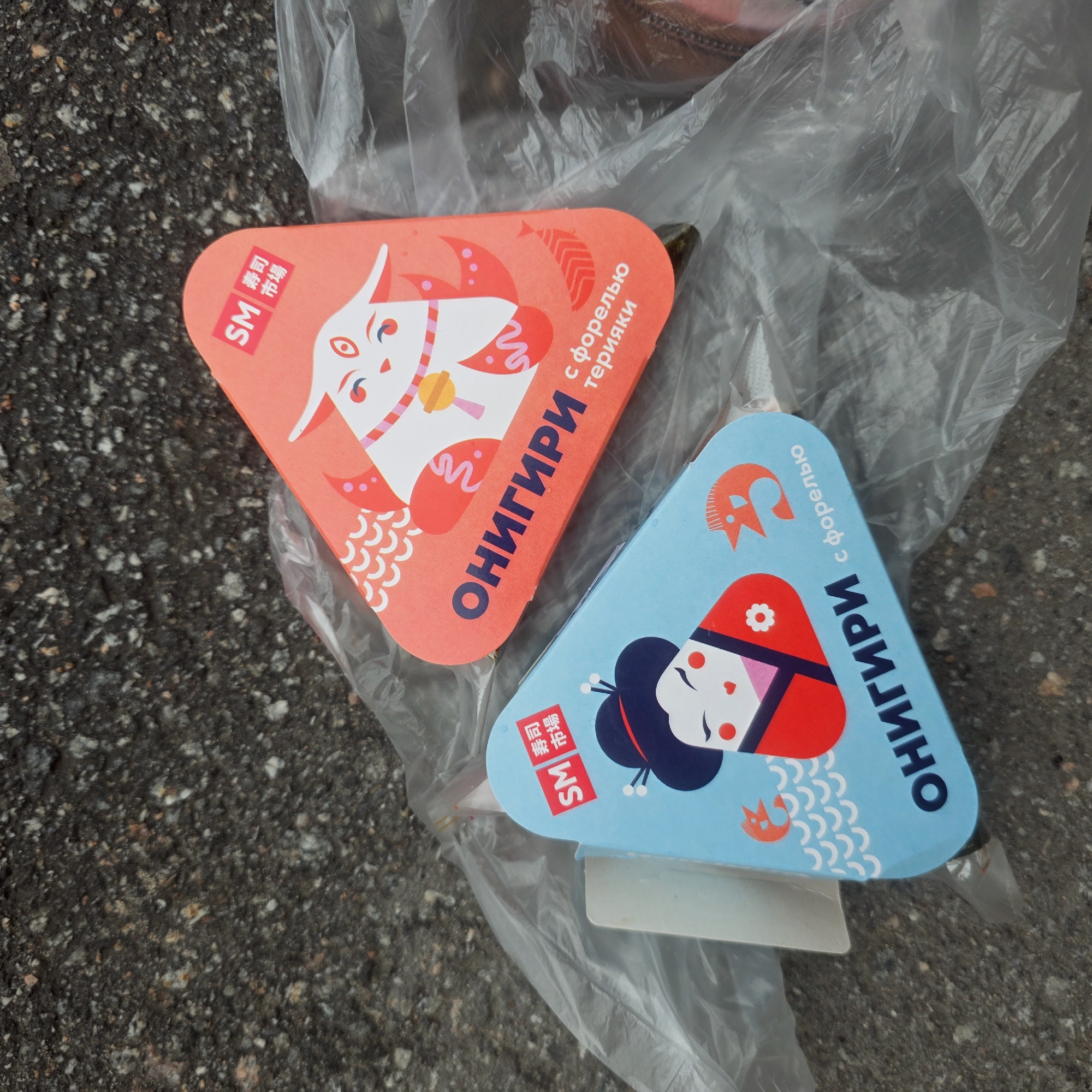 Two triangular packages of onigiri (rice balls) sit on top of a plastic bag on asphalt pavement. The onigiri are from the "SM Sushi Market" chain. The red package features a white fox illustration and the text "ОНИГИРИ с форелью терияки" ("Onigiri with trout teriyaki"). The blue package features an illustration of a girl in traditional attire and the text "ОНИГИРИ с форелью" ("Onigiri with trout"). The plastic bag is partially transparent, and some of its contents are visible. The location is likely an urban area, given the asphalt pavement. Since the packaging is in Russian, the location is likely within Russia, specifically in St. Petersburg, Russia given the brand name.