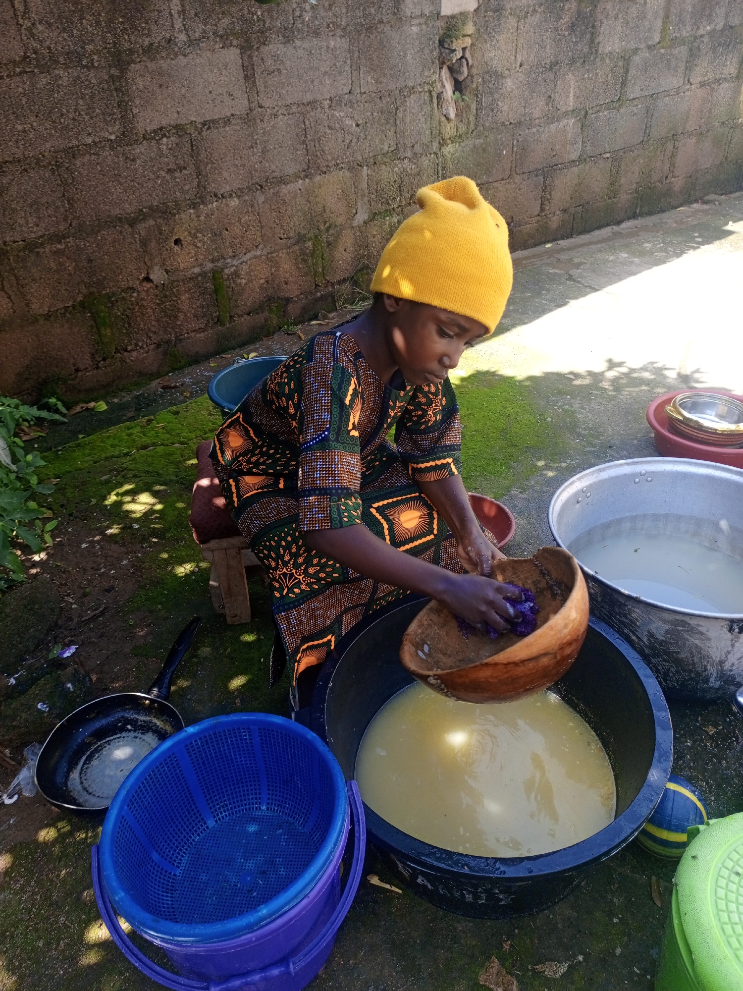 A young person wearing a yellow beanie and a vibrantly patterned dashiki is crouched on a small wooden stool. They are outdoors, in what appears to be a courtyard or an alleyway, with a rough brick wall as a backdrop and moss-covered ground. The person is actively engaged in a task, holding a large, hollowed-out gourd in their hands. Inside the gourd, they are processing a dark purple material, possibly for dyeing or preparing food. Surrounding them are various containers: a large black tub filled with a light yellow liquid, a large metal basin containing clear water, a blue plastic basket, a small black frying pan, and a red dish with metal bowls stacked inside. A yellow and blue ball and a green container are also visible. The lighting suggests it is daytime, with sunlight casting shadows on the ground. The scene appears to be in a residential area, possibly in the city of Jos, Nigeria, given the architectural style of the wall and the general environment.