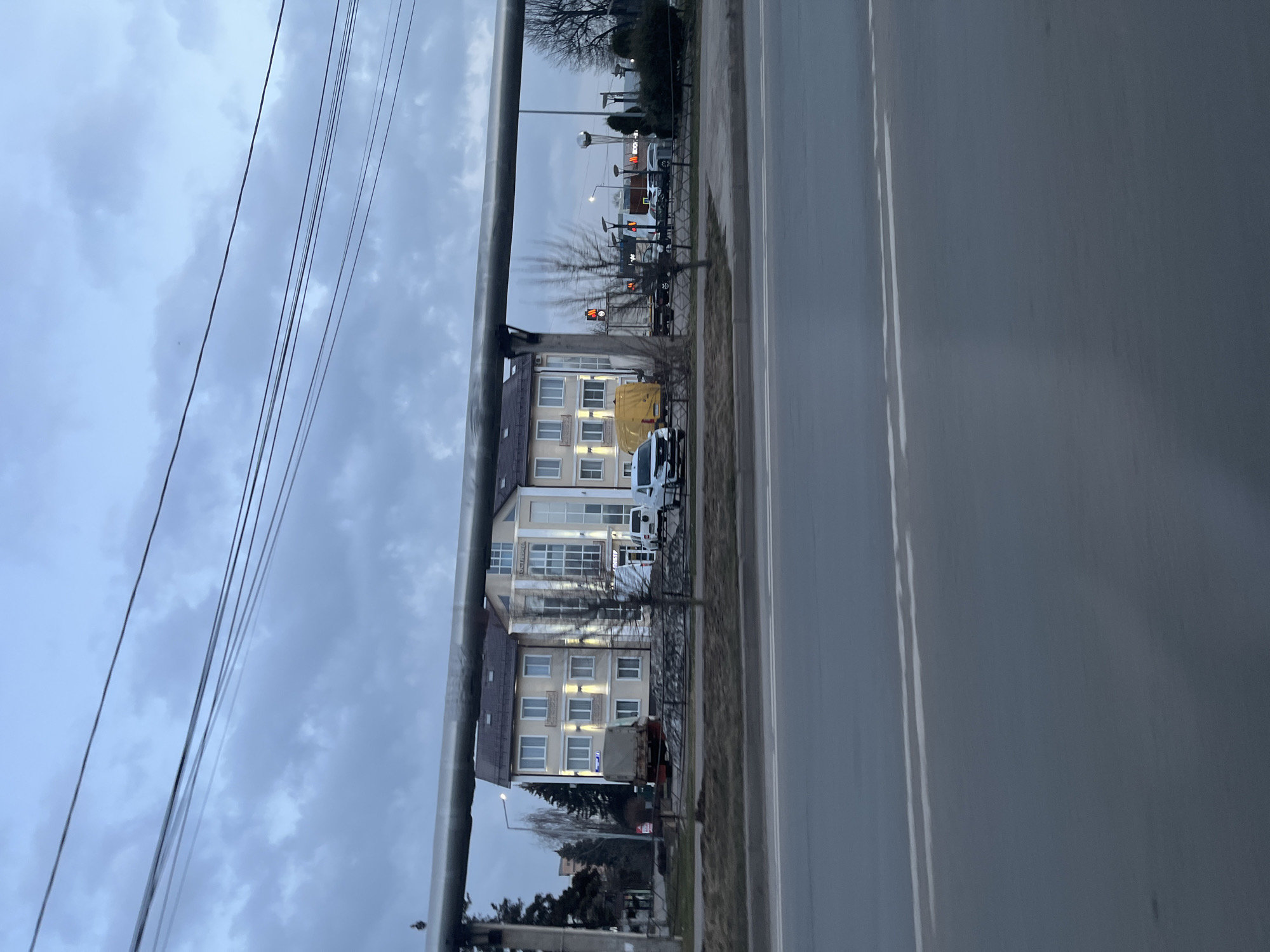 The image depicts an overcast day in Klin, Russia. A multi-story building with light-colored walls and multiple windows stands adjacent to a road. Several vehicles, including a yellow van and a white car, are parked near the building. A large metal pipe runs along the left side of the frame, with electrical wires visible above. In the distance, streetlights and a traffic signal can be seen. The road occupies a significant portion of the right side of the image.