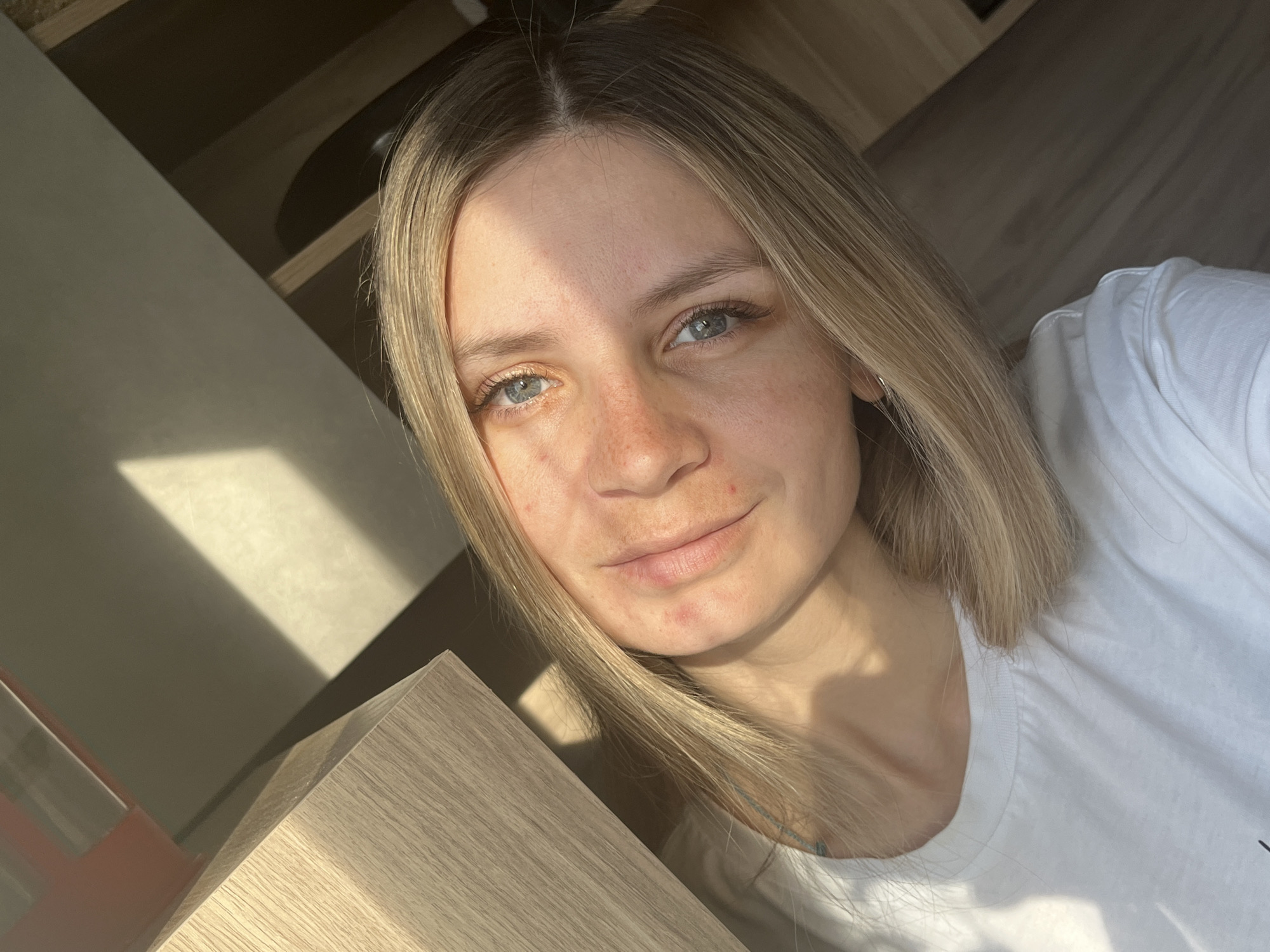 A close-up selfie of a blonde-haired woman with fair skin and blue eyes. She is wearing a white t-shirt. The background features a wooden surface and shelf. Sunlight is illuminating the scene, creating shadows on the woman's face and surrounding surfaces. No specific details link the image to Chaikovsky, Russia.