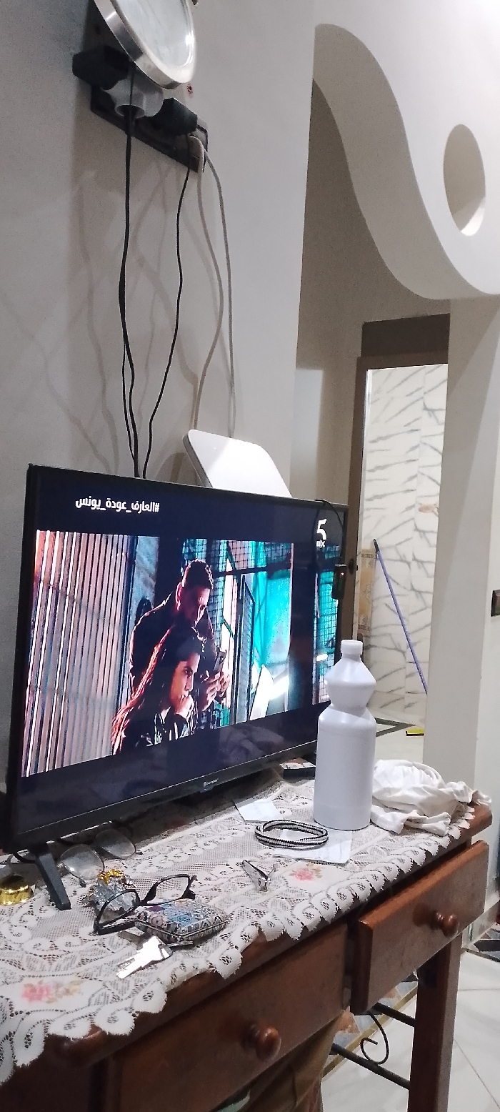 This image captures an interior residential scene in Aïn El Aouda, Morocco, depicting a television displaying a program, surrounded by various household objects on a wooden table and against a cream-colored wall.
The central focus is a dark flat-screen television positioned on a brown wooden table covered by a white lace-patterned tablecloth with subtle floral motifs. The television displays a scene featuring two individuals: a male figure in a dark garment, looking over the shoulder of a female figure with dark hair, both intently focused on a mobile device held by the female. The background of the TV image appears to be an urban or industrial setting with metal grates. Text in Arabic, "#العارف_عودة_يونس" (The Knower/Expert_Return_Younes), is visible in the top left corner of the screen, and a "5 mbc" channel logo is in the top right.
On the table to the right of the television, a tall, white plastic bottle with a multi-faceted design stands upright, next to a crumpled piece of white fabric. To the left of the television, a pair of black-framed eyeglasses rests near a small, ornate, multi-colored square box. A silver-colored key lies on the tablecloth, and a black and white beaded bracelet is positioned further to the right. The wooden table features two visible drawers with circular knobs and decorative wrought-iron scrollwork underneath.
Above the television, mounted on the wall, is a circular metallic object, possibly a clock or mirror. Below it, a black power strip is visible, with several black electrical cables plugged in and draped downwards, casting shadows on the wall. A white, rectangular device, possibly a Wi-Fi router, is situated on a shelf or surface above the TV, to the right of the power strip.
To the right of the scene, a doorway reveals an adjacent room with distinct white and grey patterned tile walls. A purple-handled broom or mop leans against the far wall within this visible area. The ceiling in the primary room features a decorative curved design with a circular cut-out. The overall environment suggests a lived-in domestic setting.