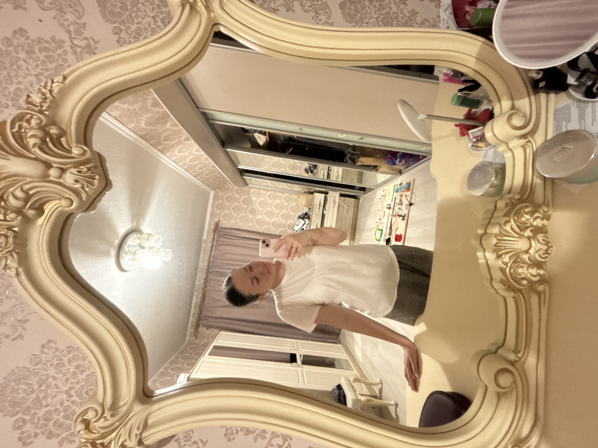 A mirror reflection reveals a woman taking a selfie in a room, likely a bedroom, given the presence of a wardrobe and dresser. The room has patterned wallpaper and a decorative ceiling light. The mirror itself is ornate, with an antique style frame. An assortment of personal items are visible on a nearby surface next to the mirror. The location is noted as Ферма, Russia.