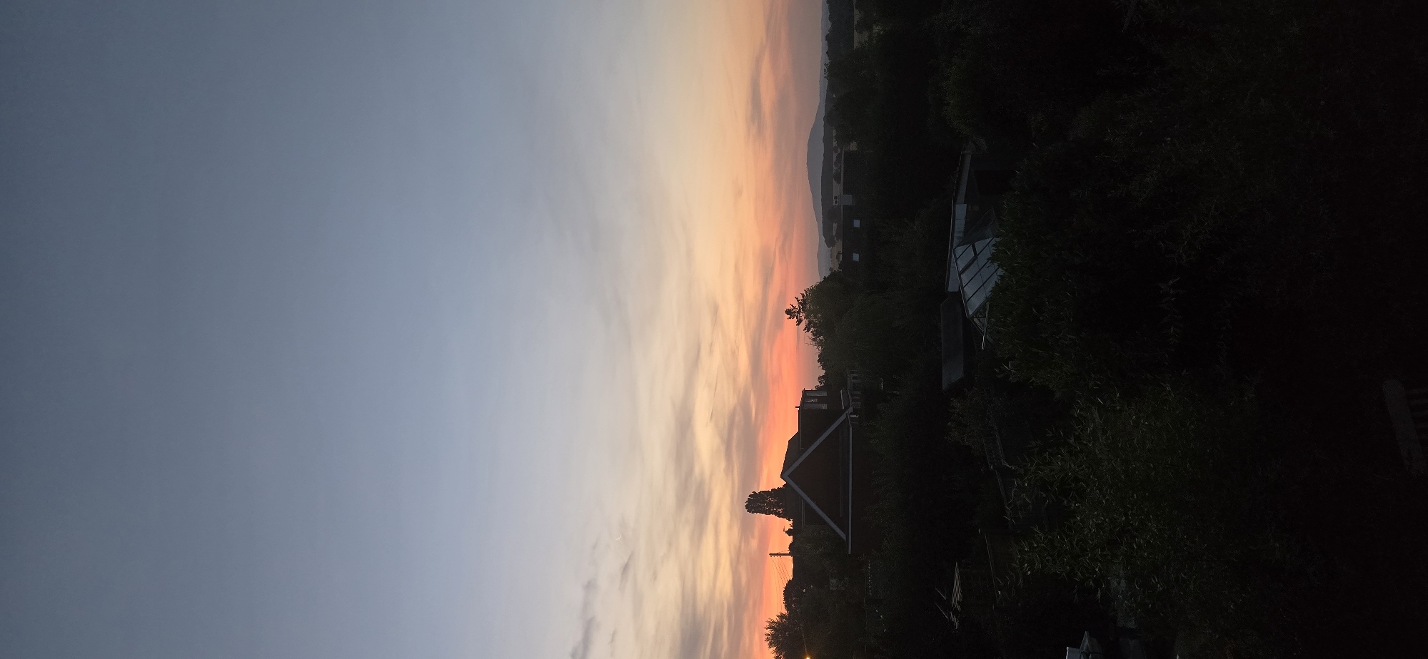 A vertical, full shot captures a twilight scene above Bayston Hill, United Kingdom. The sky transitions from a pale blue at the top to a warm orange and pink hue near the horizon, streaked with wispy clouds. Silhouetted against the vibrant sky are the dark shapes of trees, a red-roofed house with a prominent gable, and distant hills. In the foreground, dense, dark foliage obscures much of the ground, with hints of structures like a greenhouse visible within the shadows. The overall impression is one of a tranquil evening in a suburban or semi-rural setting.