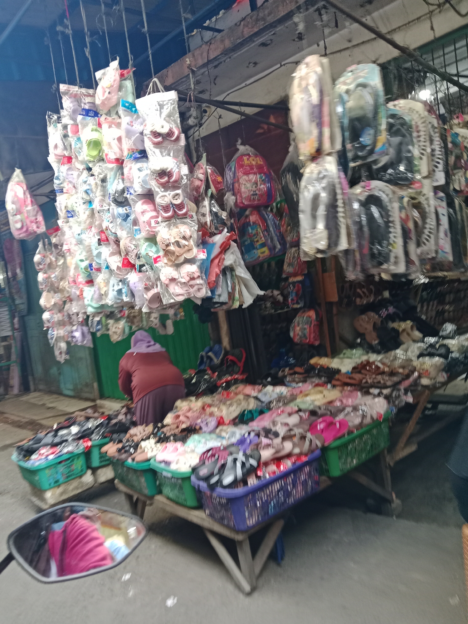 The image depicts a street-level view of a market stall in the City of Binjai, Indonesia. The stall is laden with various footwear items, predominantly children's shoes and sandals, displayed in clear plastic packaging and hanging from racks. Several colorful backpacks are also visible, along with other merchandise bagged for sale. In the foreground, a person wearing a purple headscarf and a maroon top is tending to the merchandise spread across the ground, suggesting they are a vendor. The market appears to be outdoors, with a backdrop of what looks like a green corrugated metal wall and wooden structures. The overall scene suggests a bustling, informal retail environment characteristic of many Indonesian street markets. The perspective is slightly blurred, indicating the photo may have been taken from a moving vehicle, possibly a motorcycle, as suggested by the reflection in a mirror visible in the lower left corner.