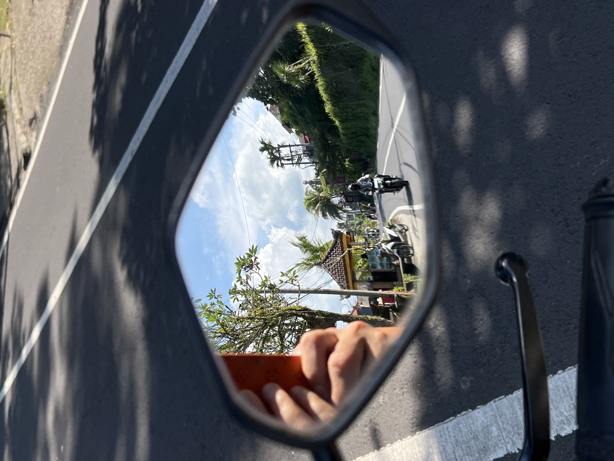 The image shows a view reflected in a motorcycle side mirror. The mirror's reflection includes a road in Petulu, Indonesia, lined with tropical vegetation and buildings. Two motorcycles are visible on the road behind the photographer. The photographer's hand, holding an orange-cased device (likely a phone), is partially visible in the lower portion of the mirror's reflection. The immediate foreground of the image shows the motorcycle's handlebars, mirror frame, and the road upon which the motorcycle is traveling. The scene is brightly lit.