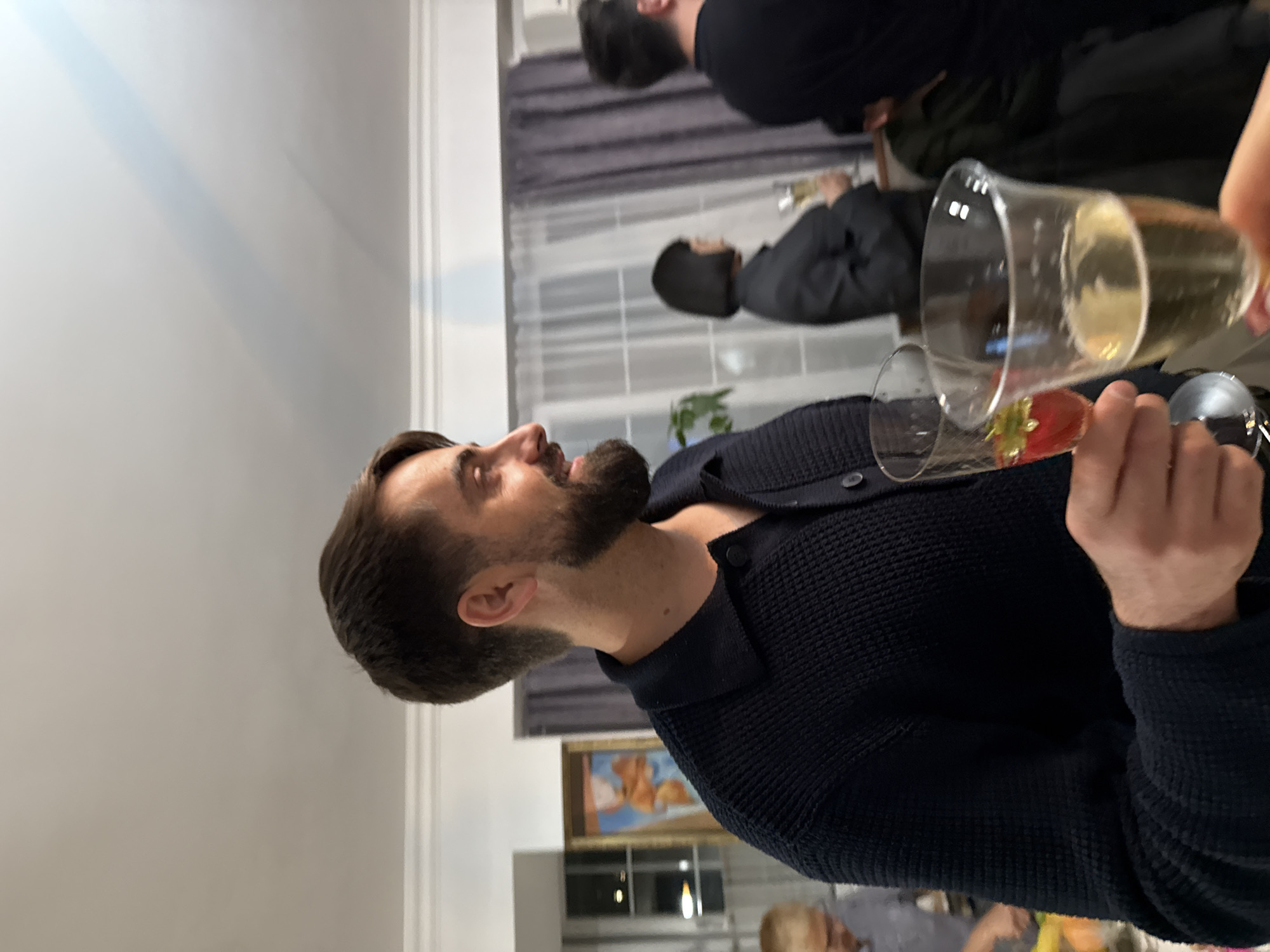 A group of people are gathered indoors, likely at a social event. One man with a beard and dark hair, wearing a navy blue cardigan, is prominently positioned in the foreground, holding a champagne glass. Another glass filled with liquid and a strawberry can be seen beside him. Background figures are partially obscured, suggesting a crowded atmosphere. A curtained window is visible in the middle ground. The presence of art on the wall implies the setting is possibly a private residence, but there is insufficient information to place the scene in the city of Yerevan, Armenia.