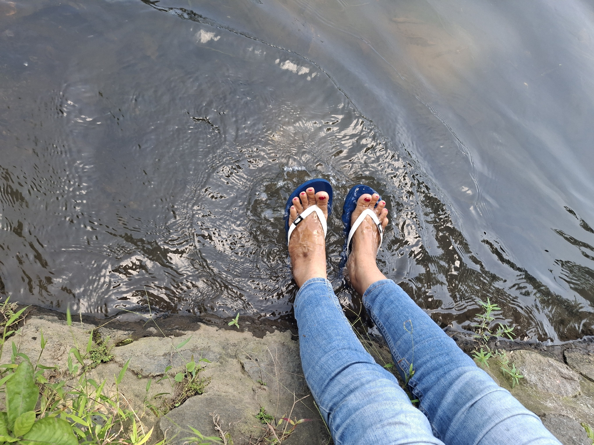 A top-down, high-angle shot shows a person's legs and feet submerged in dark, rippling water next to a rocky embankment with sparse green vegetation. The person is wearing blue jeans and blue flip-flops with white straps. Their toes, painted with red nail polish, are visible above the waterline. The water's surface is disturbed by ripples, creating a dynamic pattern of light and shadow. The rocky bank is textured and uneven, with small plants growing in crevices. The image appears to be taken outdoors, possibly in a natural setting like a river or stream within the Jogotrunan area of Indonesia.