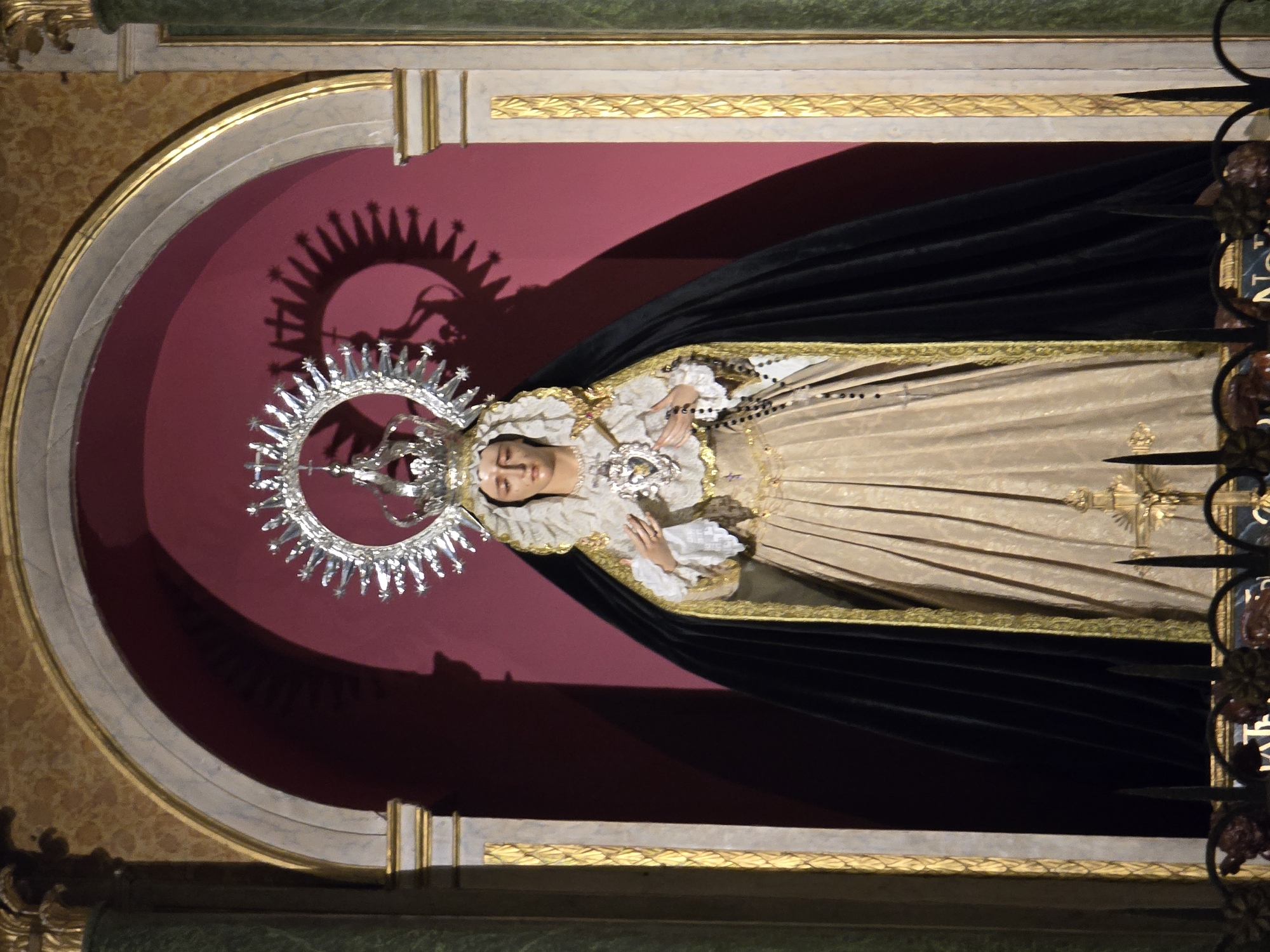 A full-shot, eye-level, interior photograph depicts a statue of the Virgin Mary, crowned and robed, within an arched alcove. The statue, situated in the city of Jaén, Spain, stands against a dark pink background, framed by columns with gold accents and a marble-like wall. The Virgin wears a radiant silver halo, a white lace-trimmed mantle, a golden pleated dress, and a black cape lined with gold. She clutches a rosary in her right hand and a heart symbol in her left. A shadow of the halo and crown is cast on the wall behind her. The bottom of the image shows a dark metal railing with decorative elements.