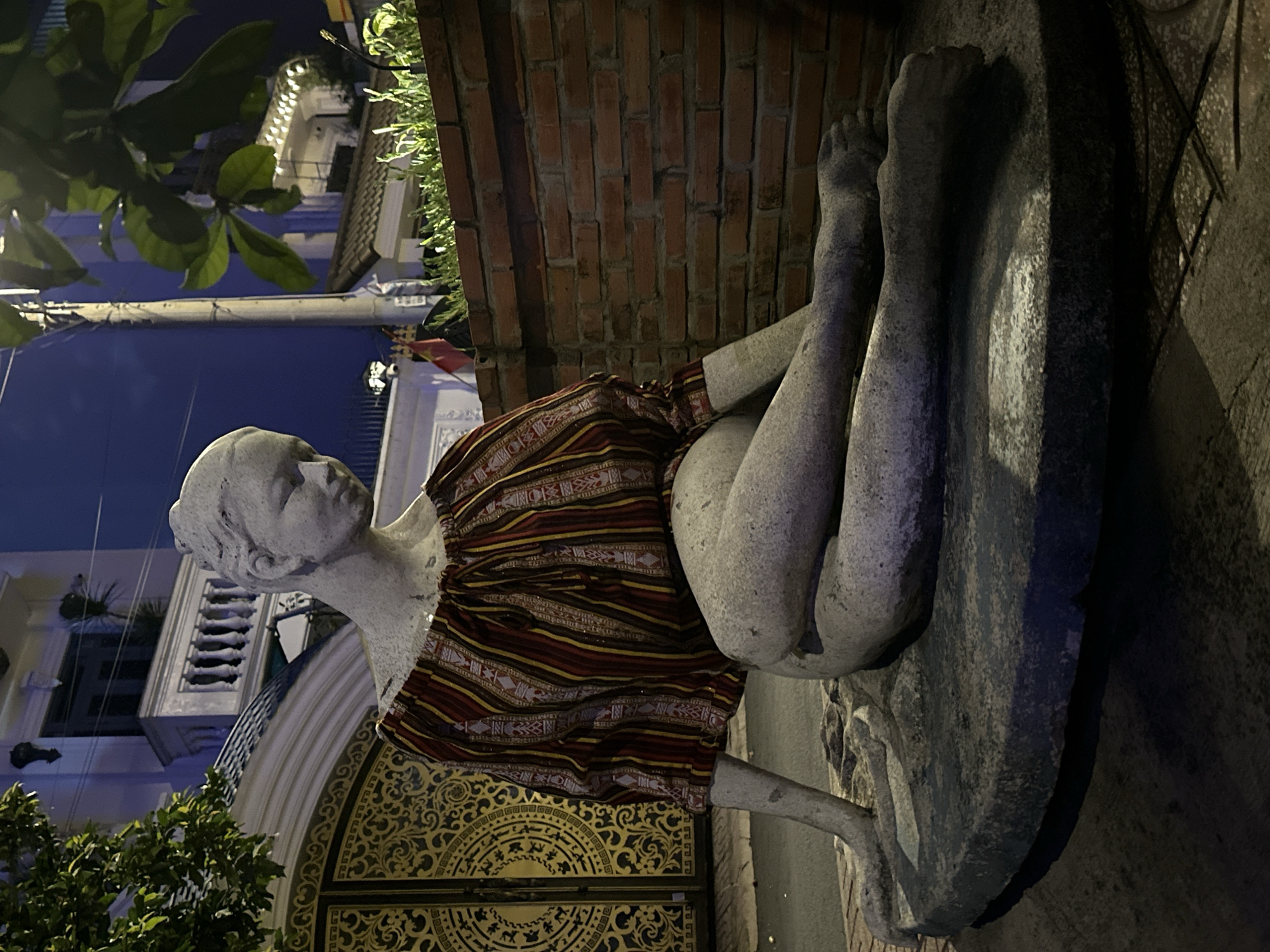 The image shows a stone statue of a reclining figure wearing a red and yellow striped garment. The statue is positioned on a stone base near a brick wall. In the background, a building with a golden ornate door and white facade can be seen. The location appears to be in a city, possibly Thủ Đức, Vietnam, based on the architecture. The lighting is dim, suggesting it is nighttime. Some trees are also visible in the frame.