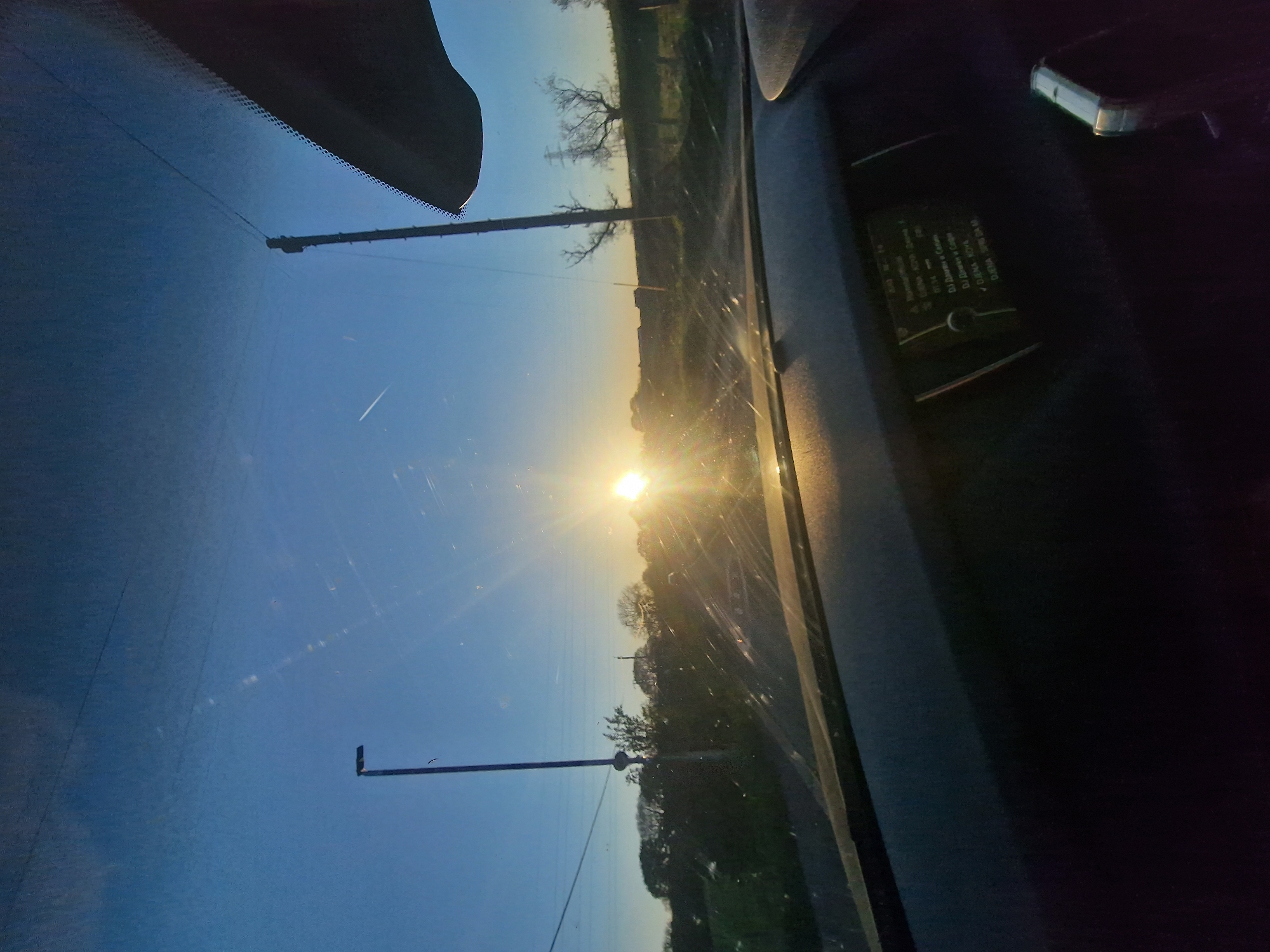 The image depicts a view through a car windshield at sunrise or sunset, seemingly taken on a road on the outskirts of Durham, United Kingdom. The sun is visible on the horizon, and power lines and foliage line the road. The car's interior is partially visible.