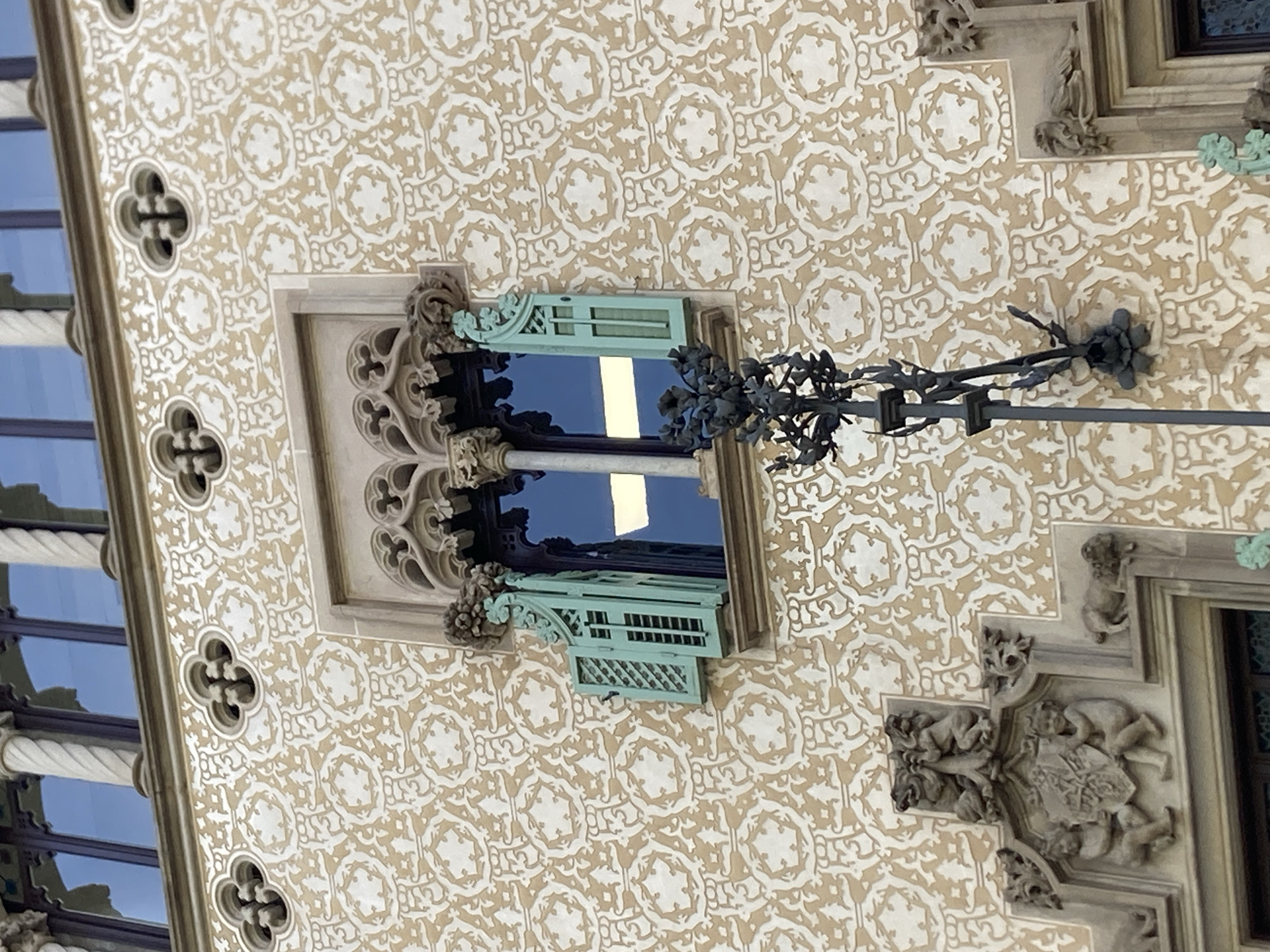 This image shows a close-up of an ornate building facade in Barcelona, Spain. The wall is decorated with a repeating pattern of stylized floral motifs in a light brown against a cream background. To the left, a section of the building features tall, narrow windows with a glass facade, reflecting a blue sky.
A prominent Gothic-style window with intricate stone tracery is centrally located. The window frame is made of stone, carved with decorative elements and figures. Inside the window, a bright reflection obscures the view. Above the window, decorative stone sculptures of mythical creatures or gargoyles are visible. To the right of the window, a decorative black metal railing with floral embellishments extends outwards. A small, light blue louvered shutter is open on the window, adding a touch of color.
The overall impression is one of intricate architectural detail and historical artistry, typical of Barcelona's rich architectural heritage. The lighting suggests it is daytime, with clear weather indicated by the bright reflections in the windows. No text is visible in the image.