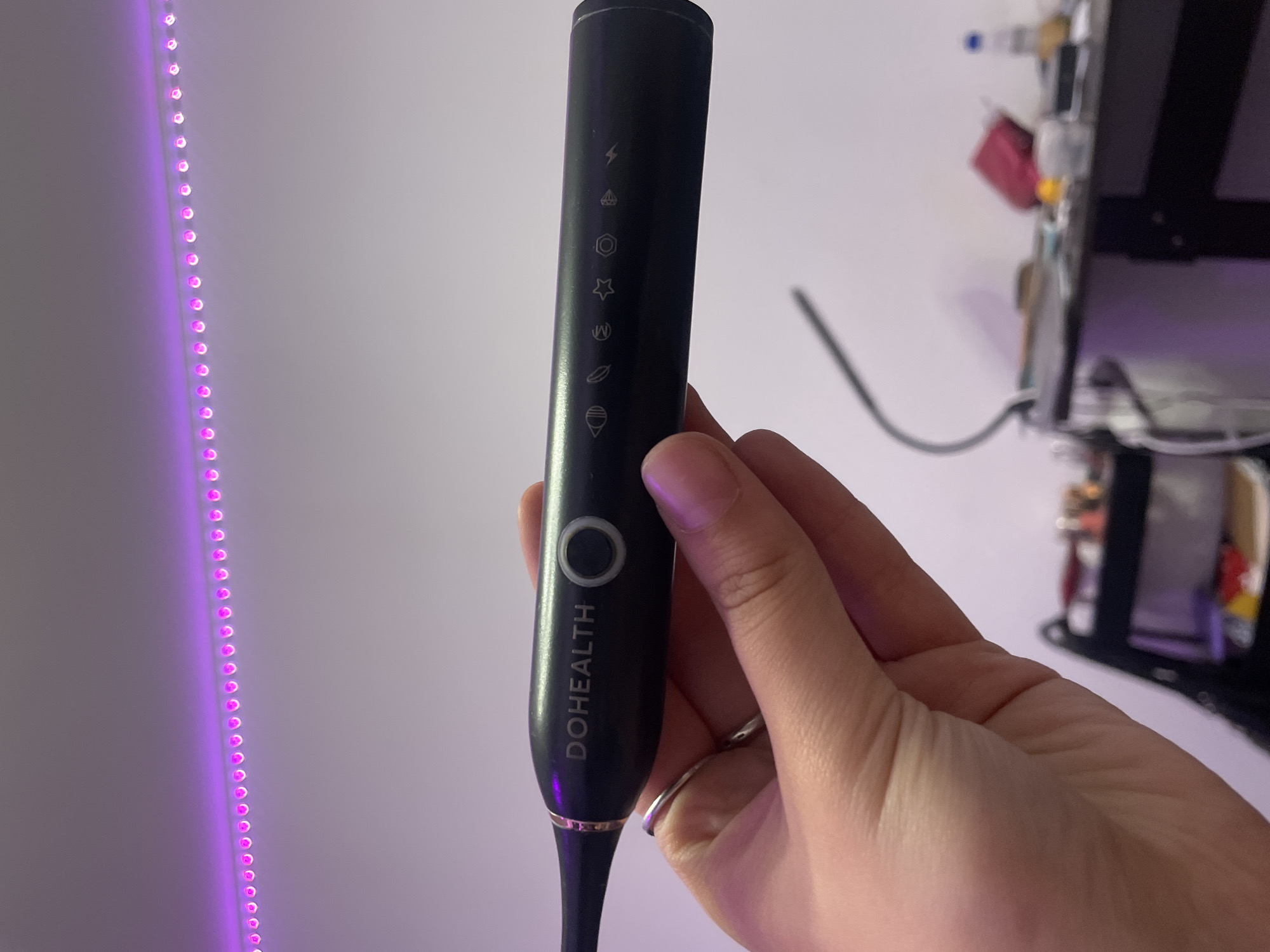 A person's hand, wearing a silver ring, holds a black electric toothbrush with "DOHEALTH" and a circular button printed on it. The toothbrush has small icons along its length, including what appears to be an eye, a leaf, a crescent moon, a star, and a lightning bolt. The background is a plain white wall illuminated by a string of pink LED lights running along the top. To the right, a blurred desk with various items is visible, including a water bottle and what looks like a red pouch. The image does not provide any information about the city of Kokkorevo, Russia.