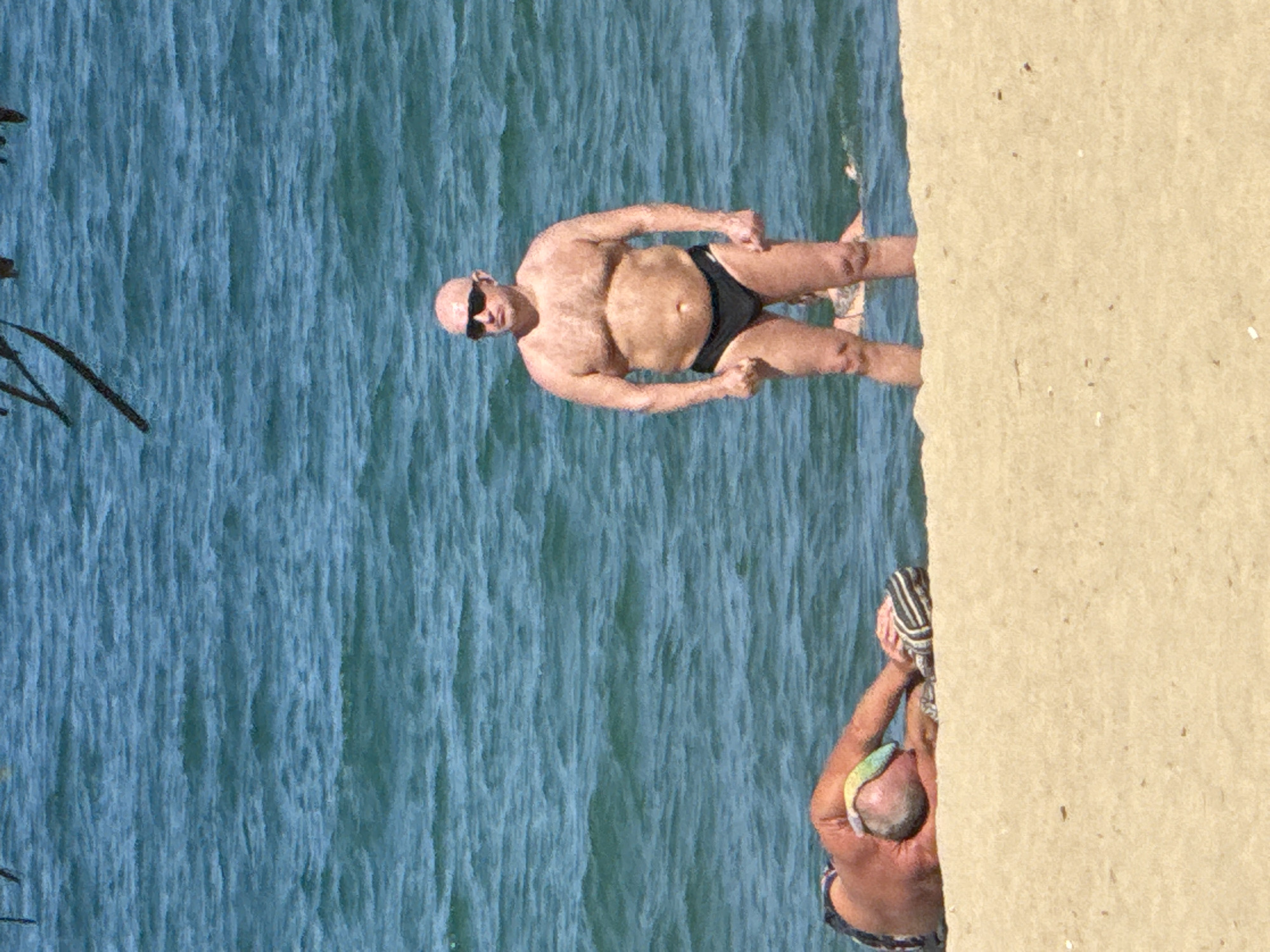 The image depicts a beach scene in Nha Trang, Vietnam, under bright daylight. The foreground consists of light tan sand. The midground and background feature blue-green ocean water with visible surface choppiness.
Two adult males are prominent. One, a bald male with a heavy build and tanned skin, stands upright in the shallow ocean water, facing slightly right of the viewer. He wears black speedo-style swim trunks and dark sunglasses, with his hands clenched at his sides. The water reaches approximately mid-thigh. A second adult male lies prone on his right side on the sand in the immediate foreground, facing left. He has gray hair and tanned skin, wearing patterned swim trunks. His head rests on a colorful striped object, and a black and white striped object is near his left hand. A third individual is partially submerged in the ocean water behind the standing male, with only their upper back and head visible, appearing to be swimming or floating. Obscured, dark, blurry linear objects are visible at the very top edge of the frame.