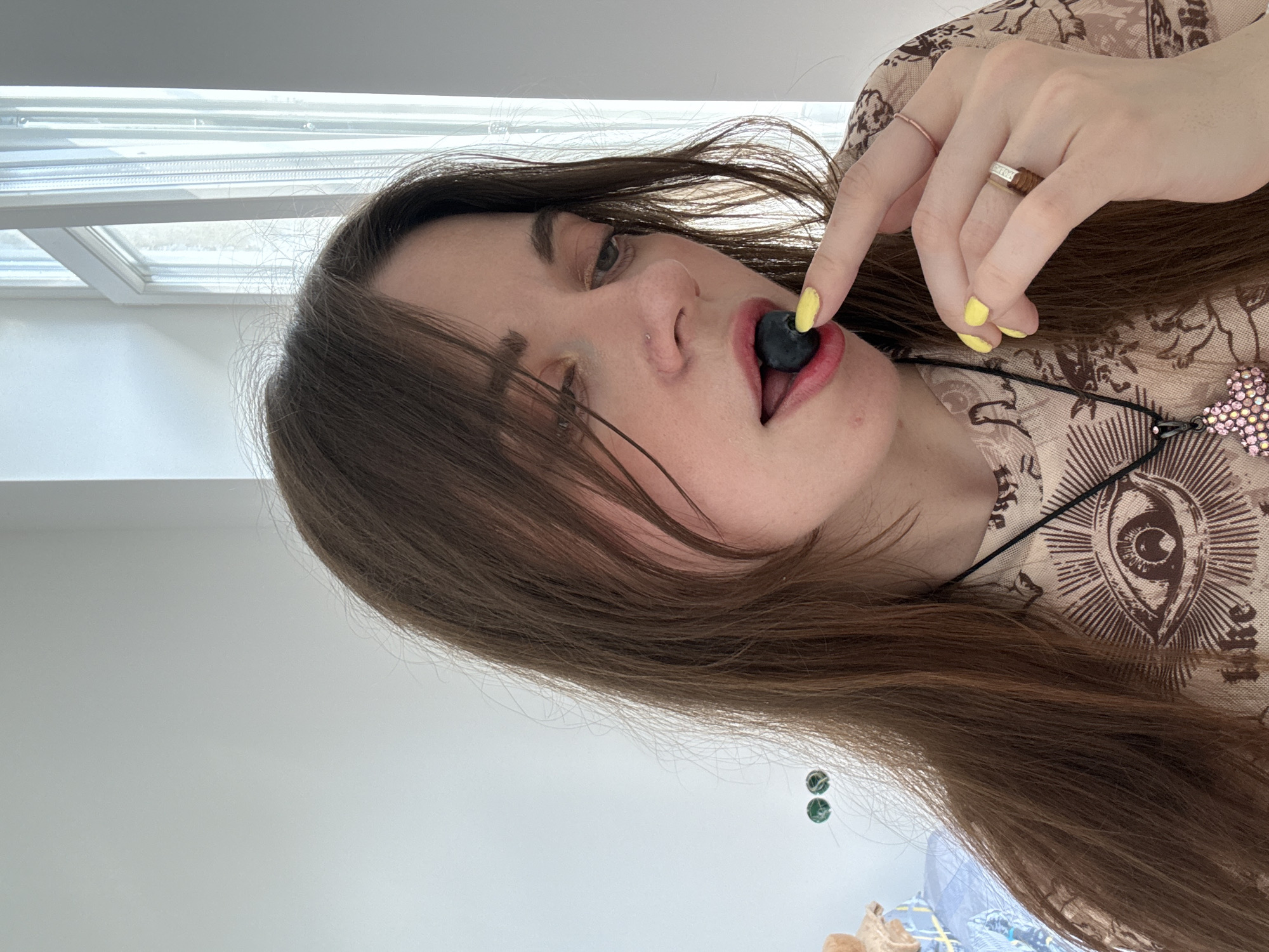 A woman with light skin and long brown hair holds a large blueberry in her mouth, positioning it with her right index finger. Her nails are painted a bright yellow. She wears two rings on the same hand, one a plain band and the other with small, inset diamonds. The woman wears a beige top with an eye print and a necklace with a decorative cross pendant. The background includes a window with blinds, a white wall, and a glimpse of bedding. The location is not specified, and there is no indication that this is Moscow, Russia.